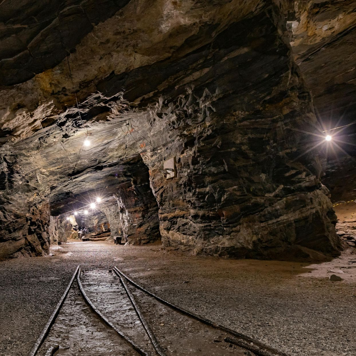 Inside of a dark mine tunnel with train tracks. Dim lights illuminate the rocky walls and floor.