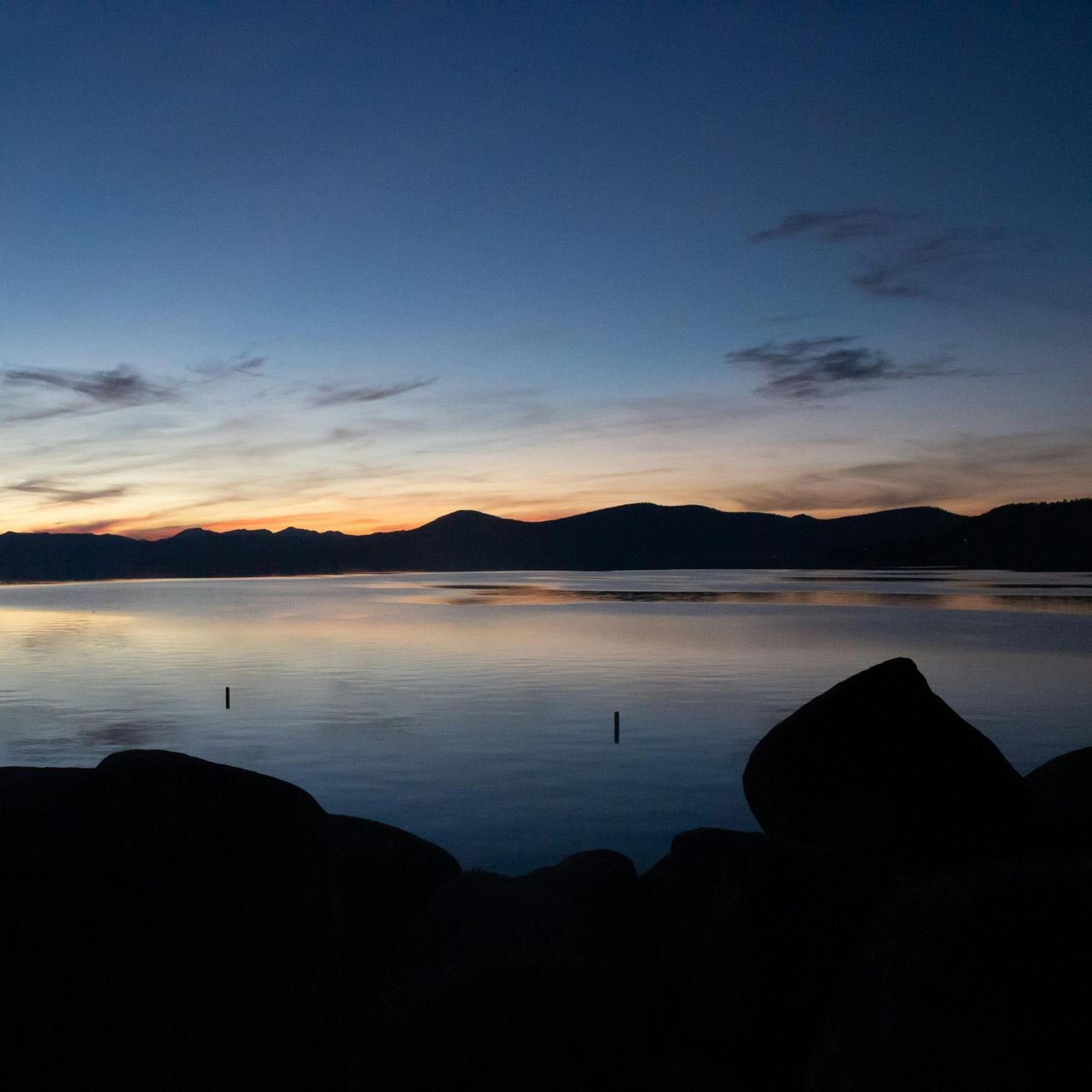 Calm lake at dusk with silhouetted mountains, rocks, and colorful sky.