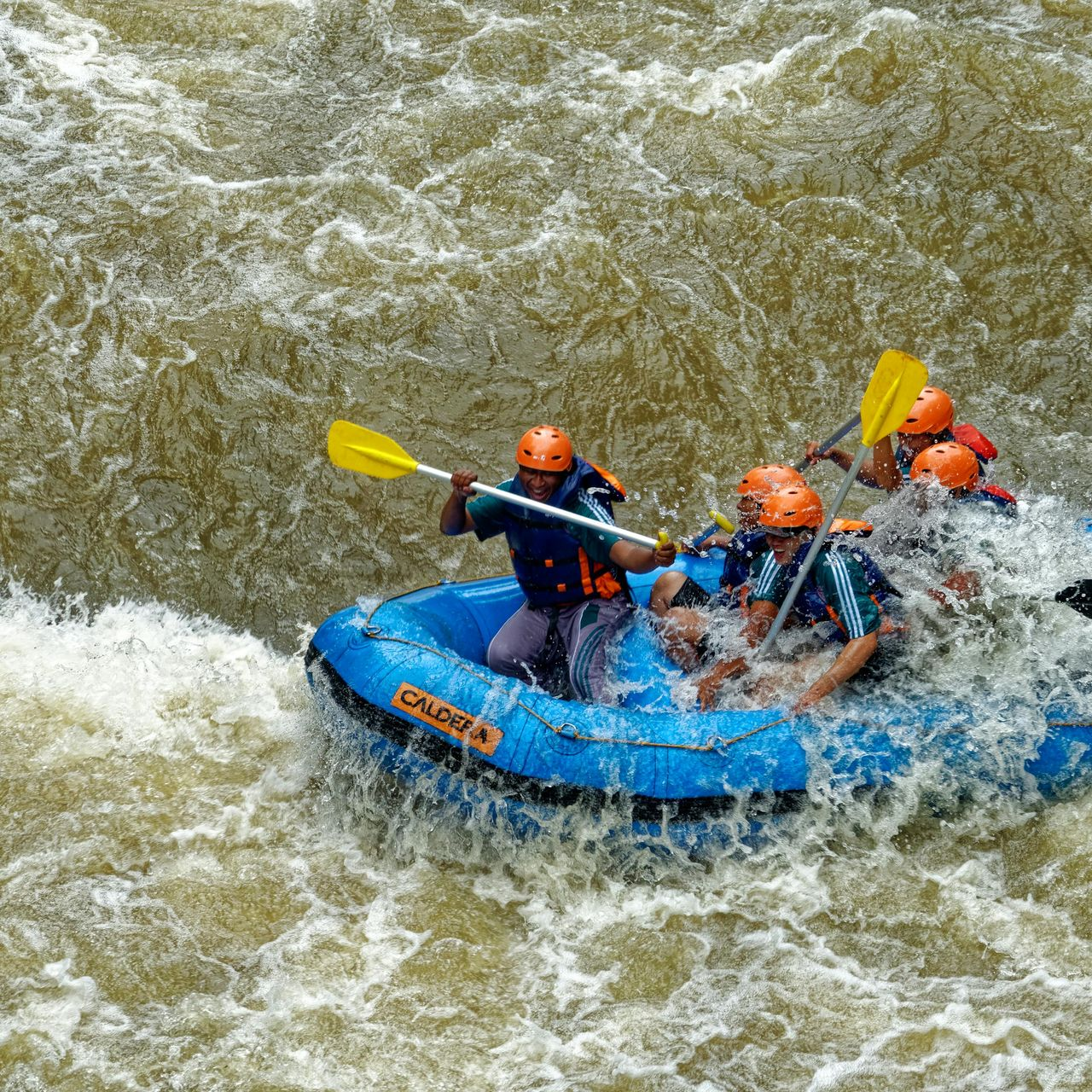 Rafting on a turbulent river; blue raft with people in orange helmets paddling through white water.
