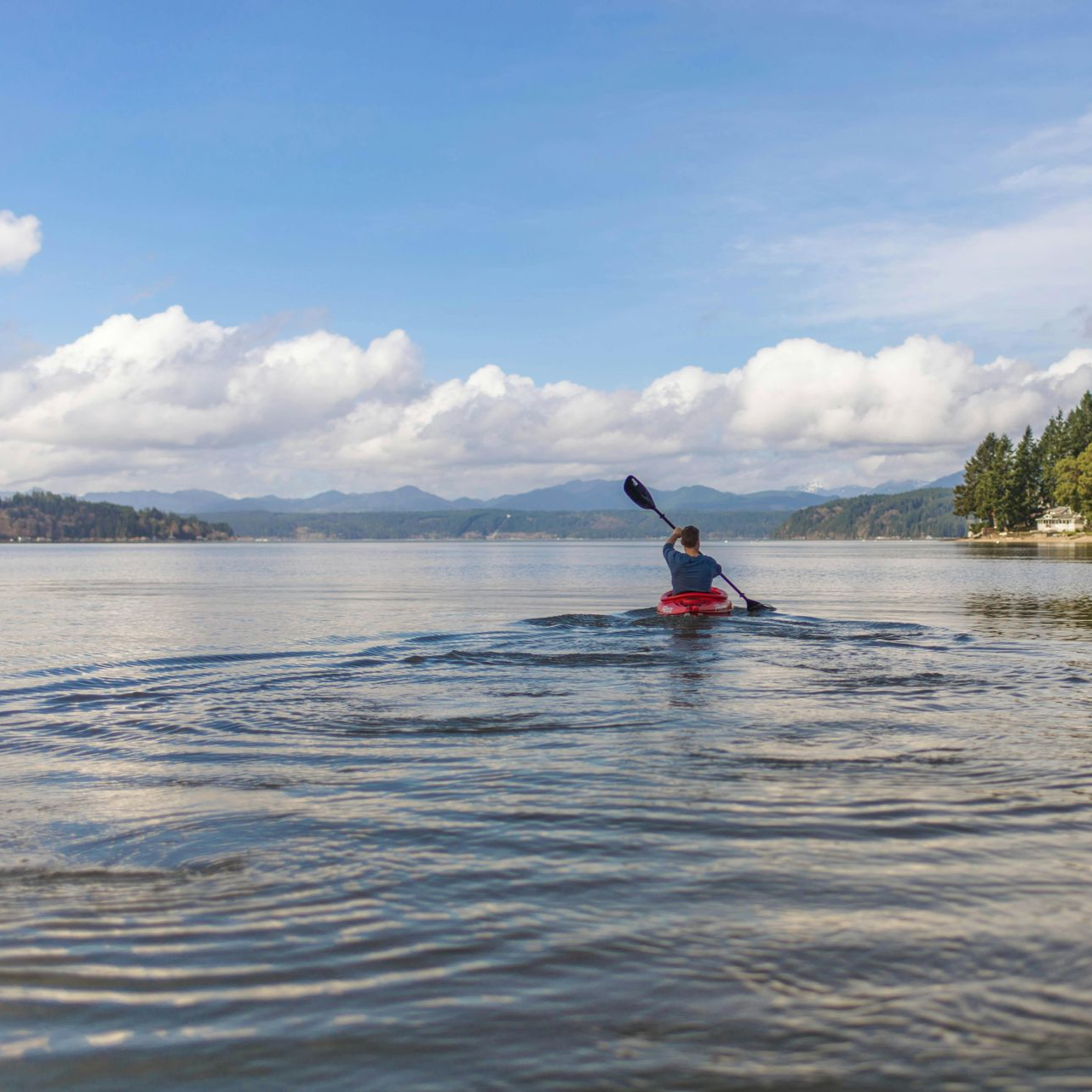 Person kayaking on calm water under a blue sky with clouds, trees in the distance.