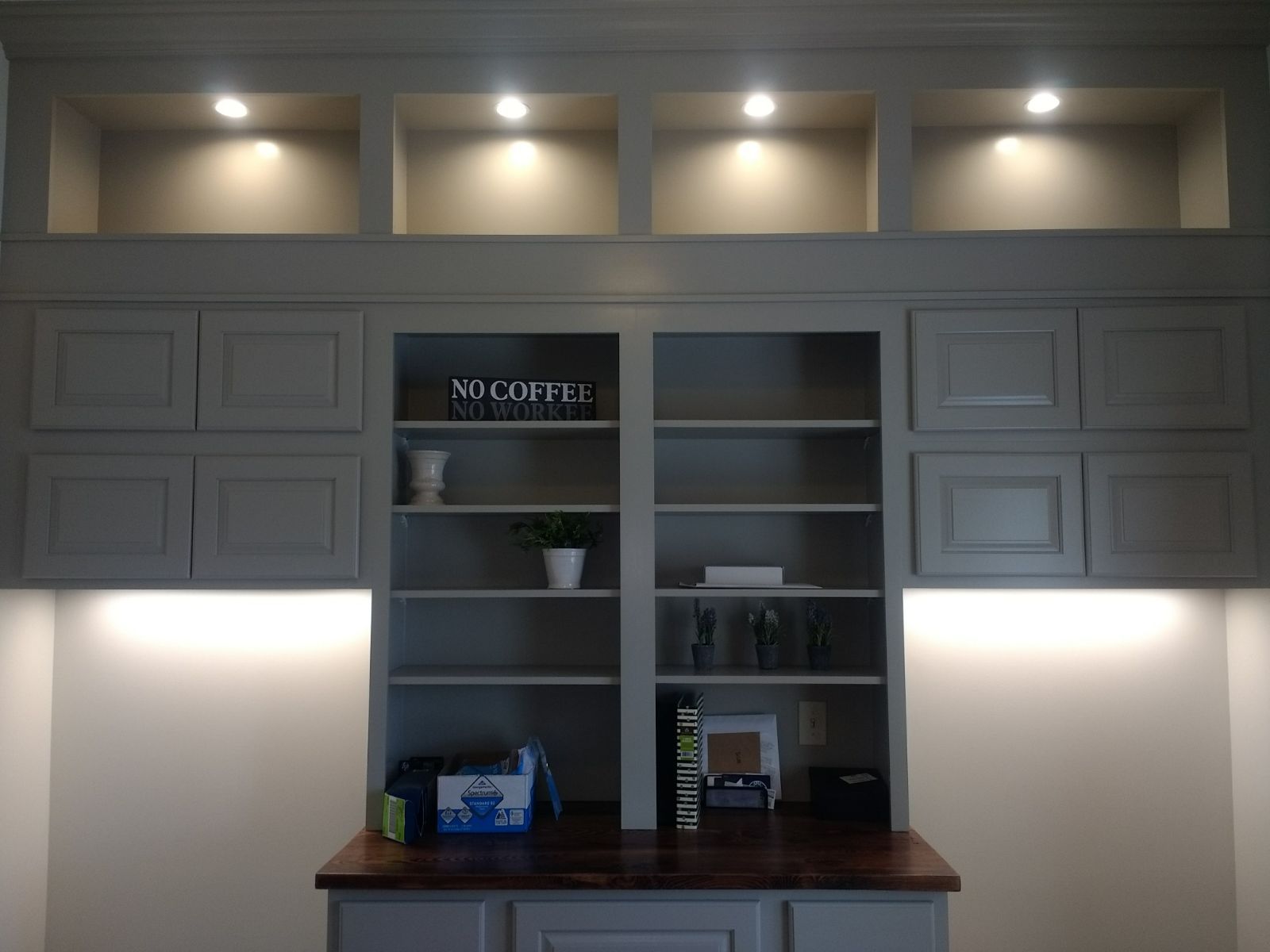 Built-in gray cabinet with shelves and upper recessed cubbies, lit with warm lights, above a dark wood desk.