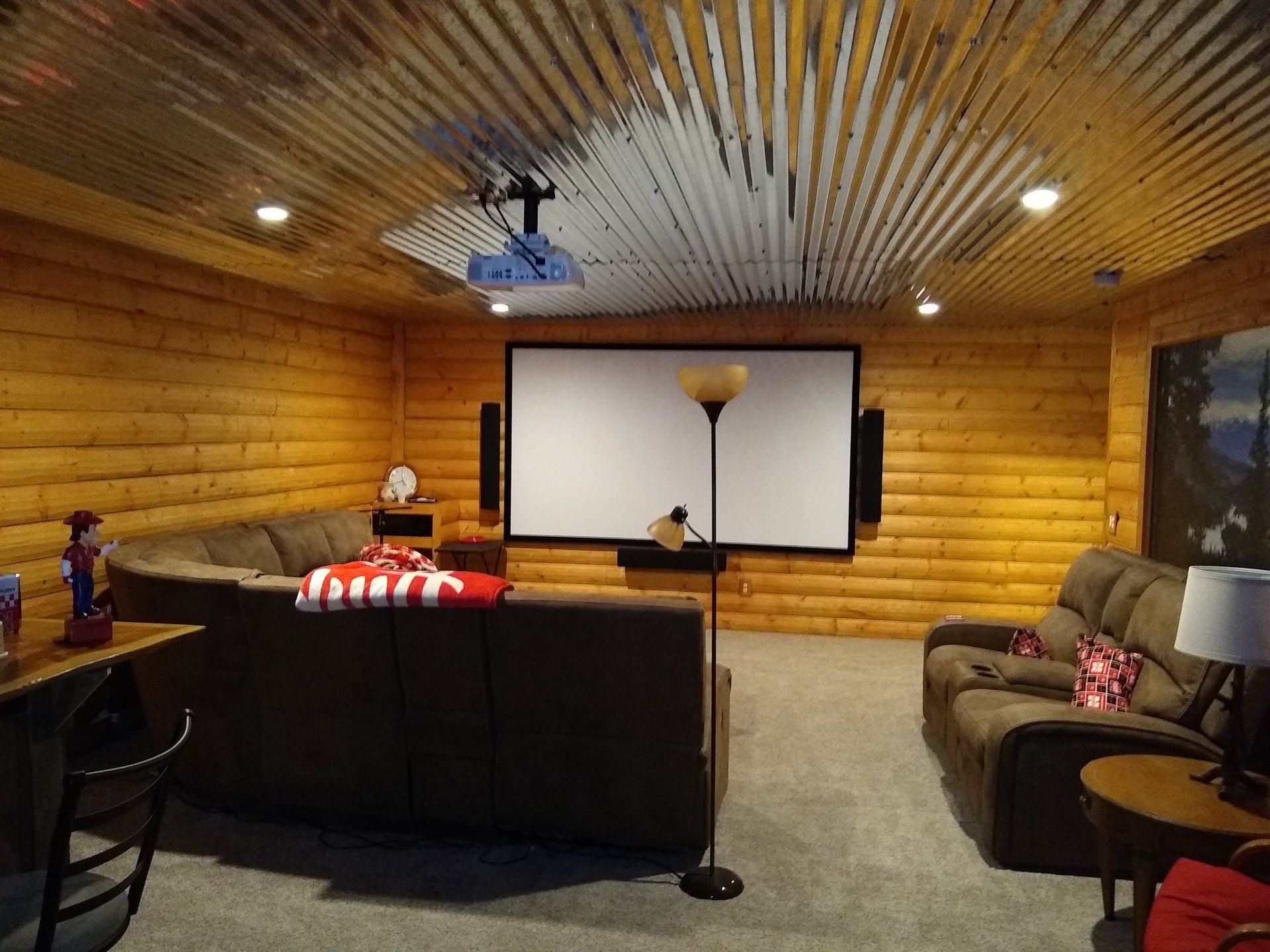 Home theater with wooden walls, metal ceiling, screen, projector, couches, and lamps.