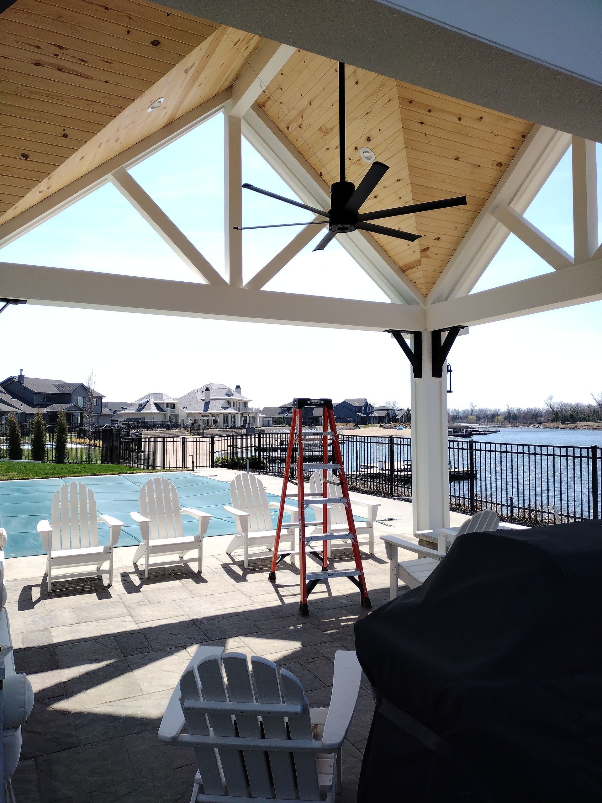 Outdoor patio with pool, water, and houses in the background, wooden ceiling, black ceiling fan, white chairs, and a red ladder.