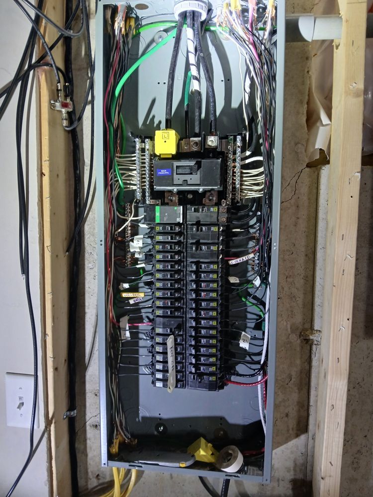 Electrical panel with numerous wires and circuit breakers inside a wall.