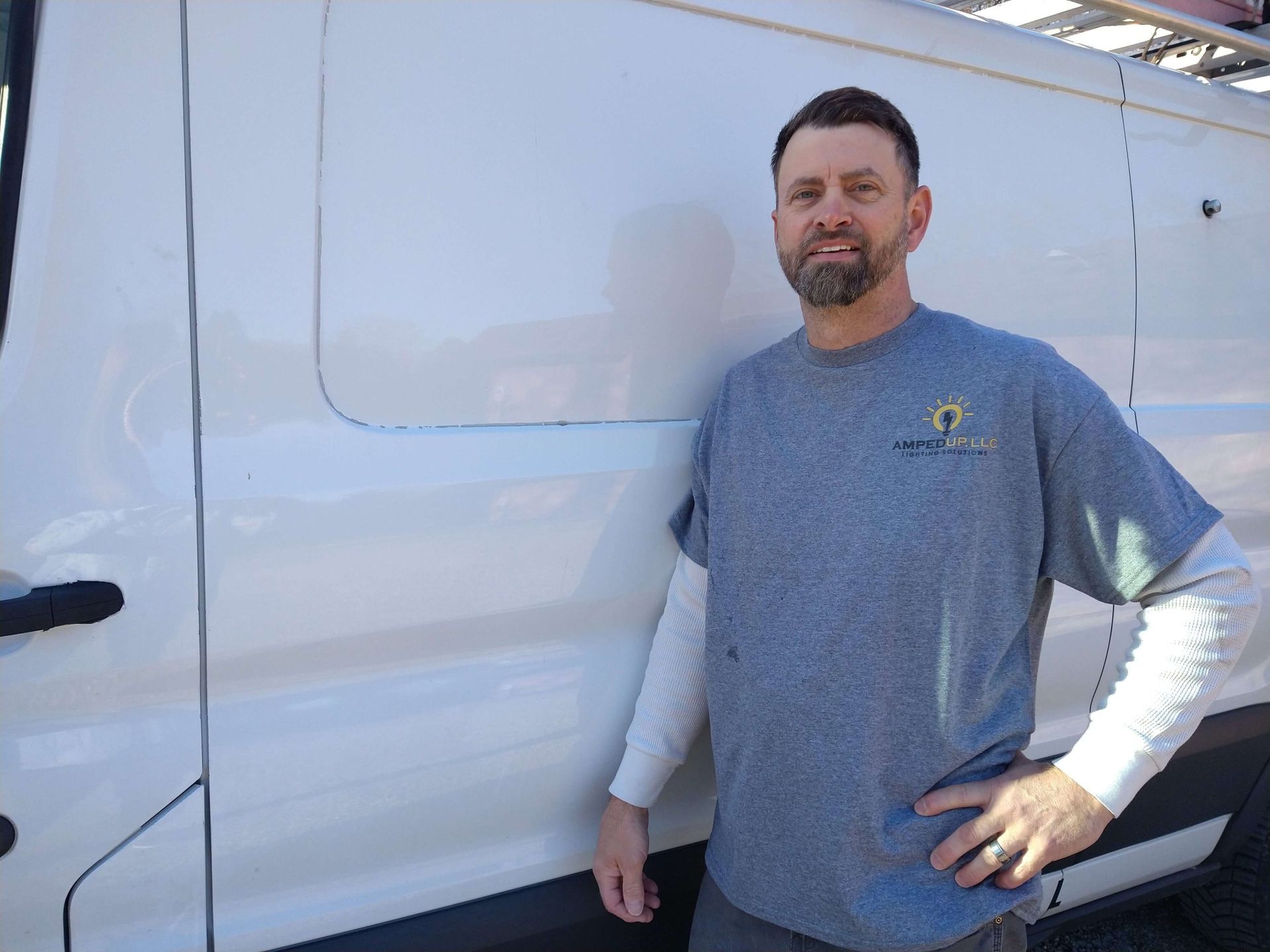 Man in gray shirt with beard, standing next to a white van with his hand on his hip, smiling.
