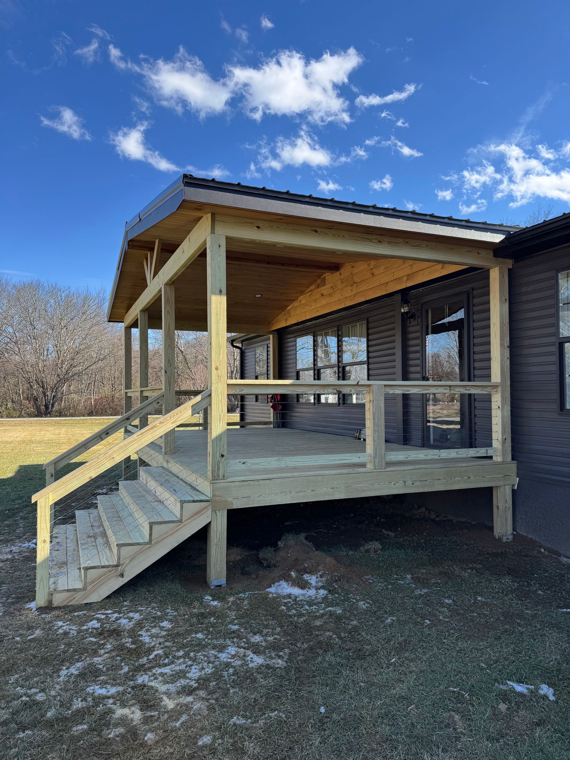 A newly constructed wooden deck and porch attached to the side of a dark-sided house under a bright blue sky.