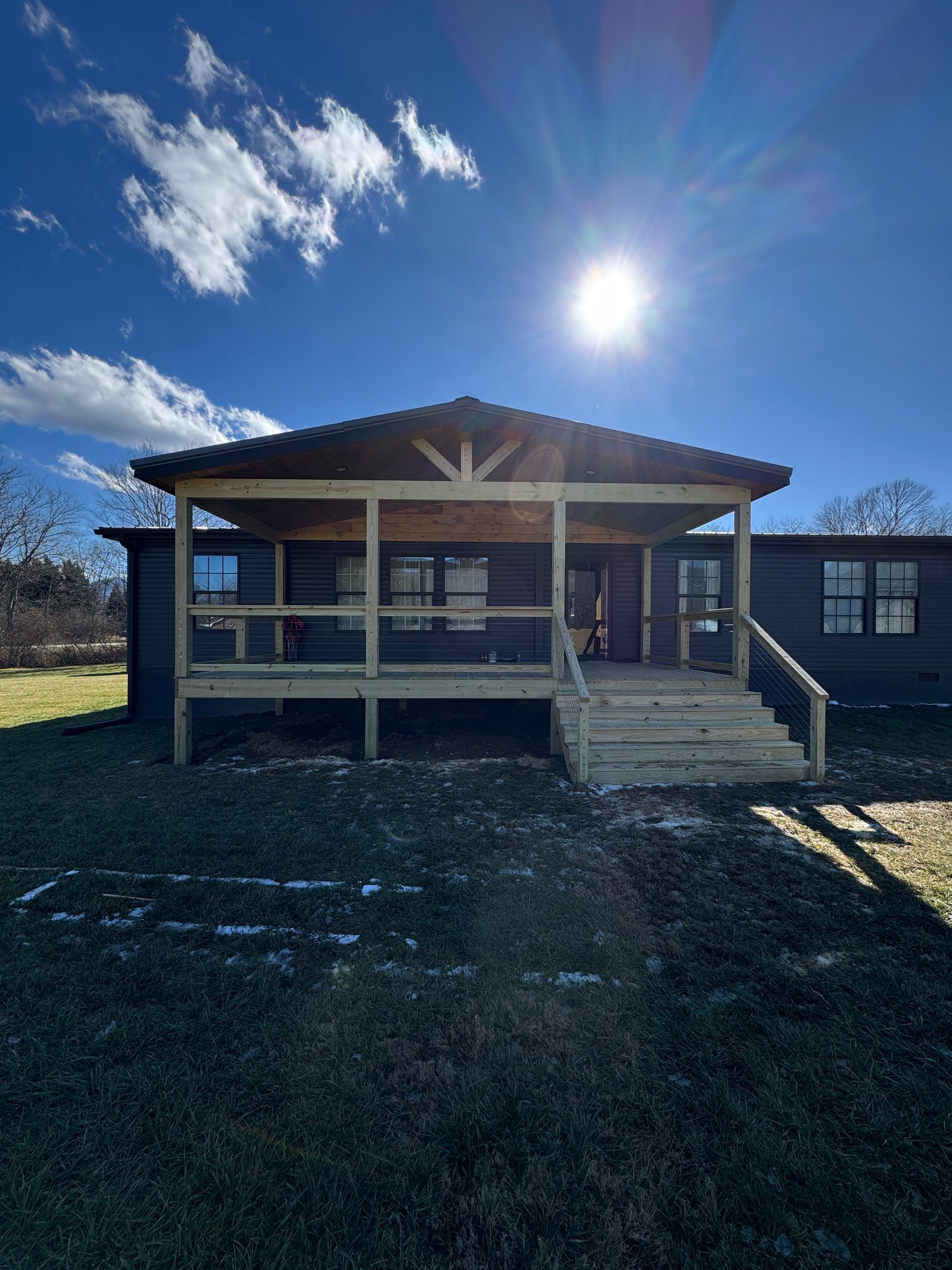 A dark-sided house with a newly constructed light-wood porch, stairs, and railing under a bright sunny blue sky.