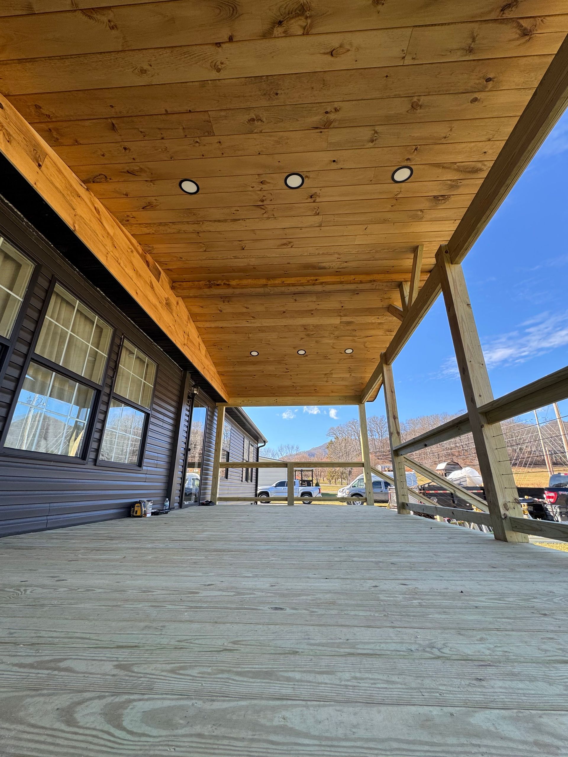 A low-angle view of a wooden porch with recessed lighting, looking out toward a clear blue sky and distant hills.