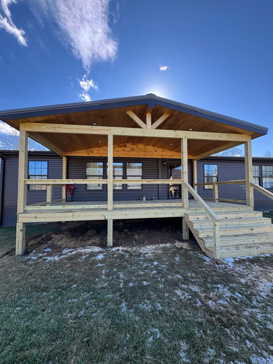 Newly constructed wooden deck with a roof overhanging a dark brown house exterior under a bright blue, sunny sky.