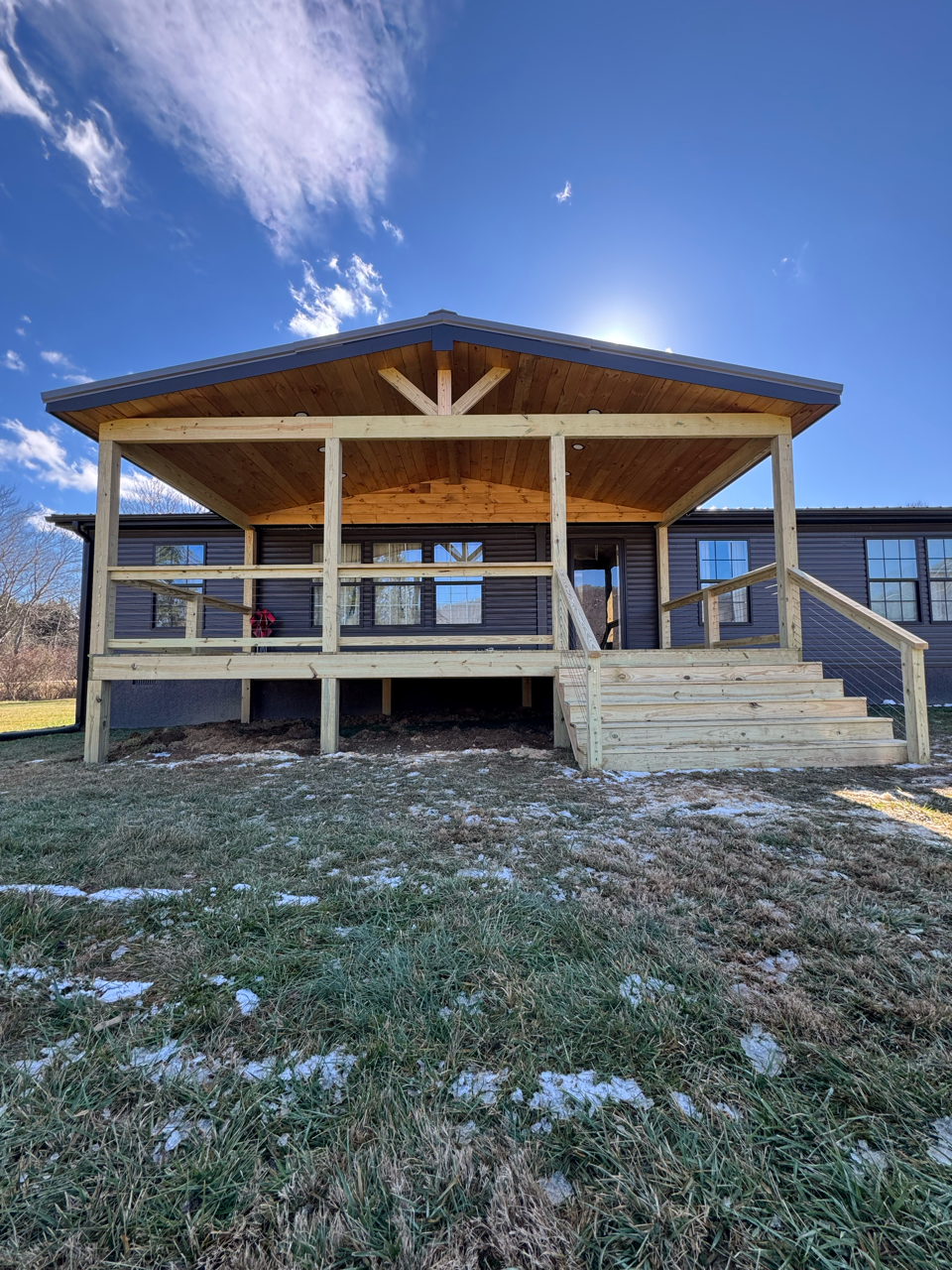 A newly built wooden deck with a covered roof attached to a dark-sided house under a bright blue sky.
