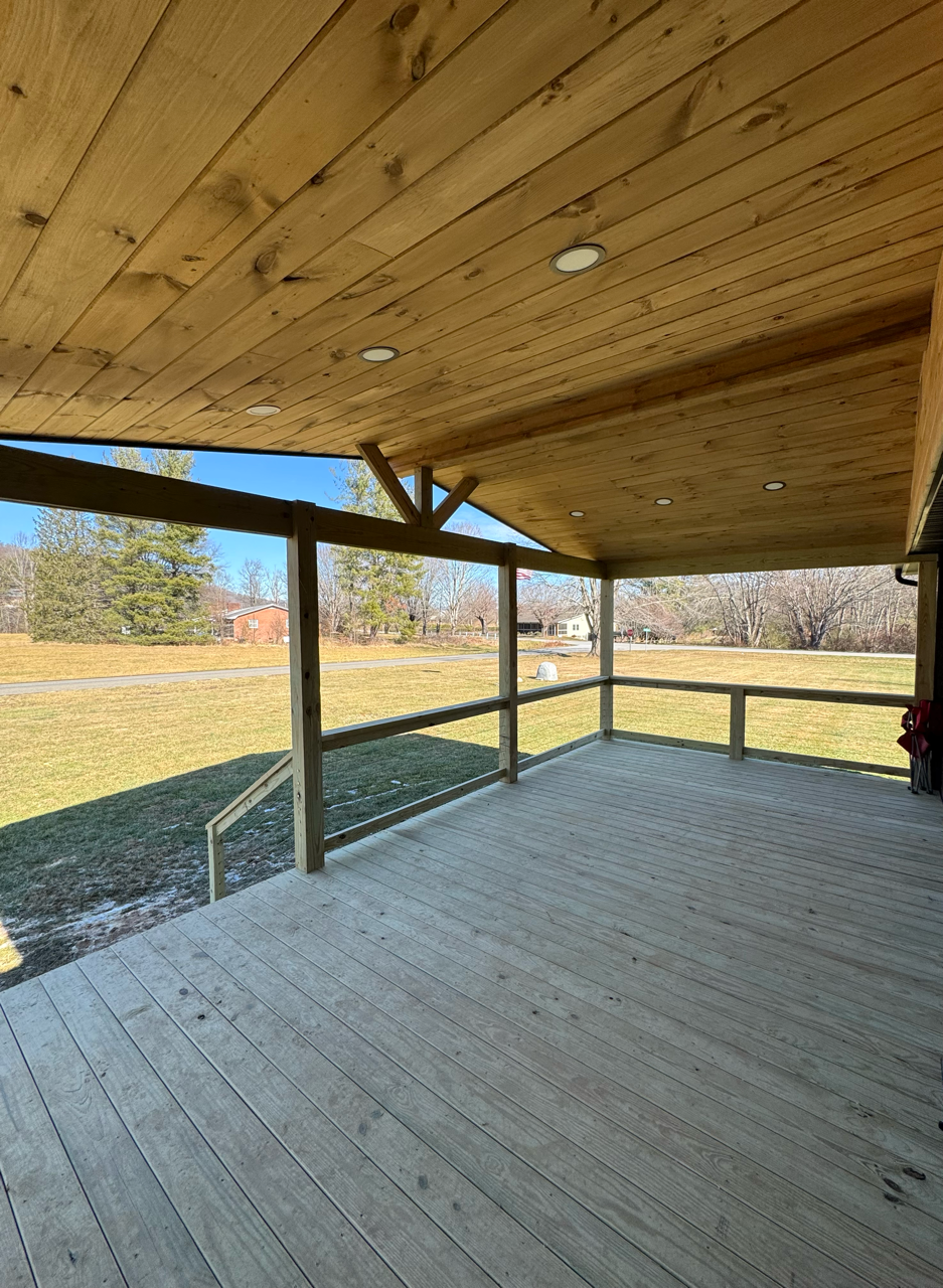A covered wooden deck with a sloped ceiling overlooking an open grassy field under a clear blue sky.