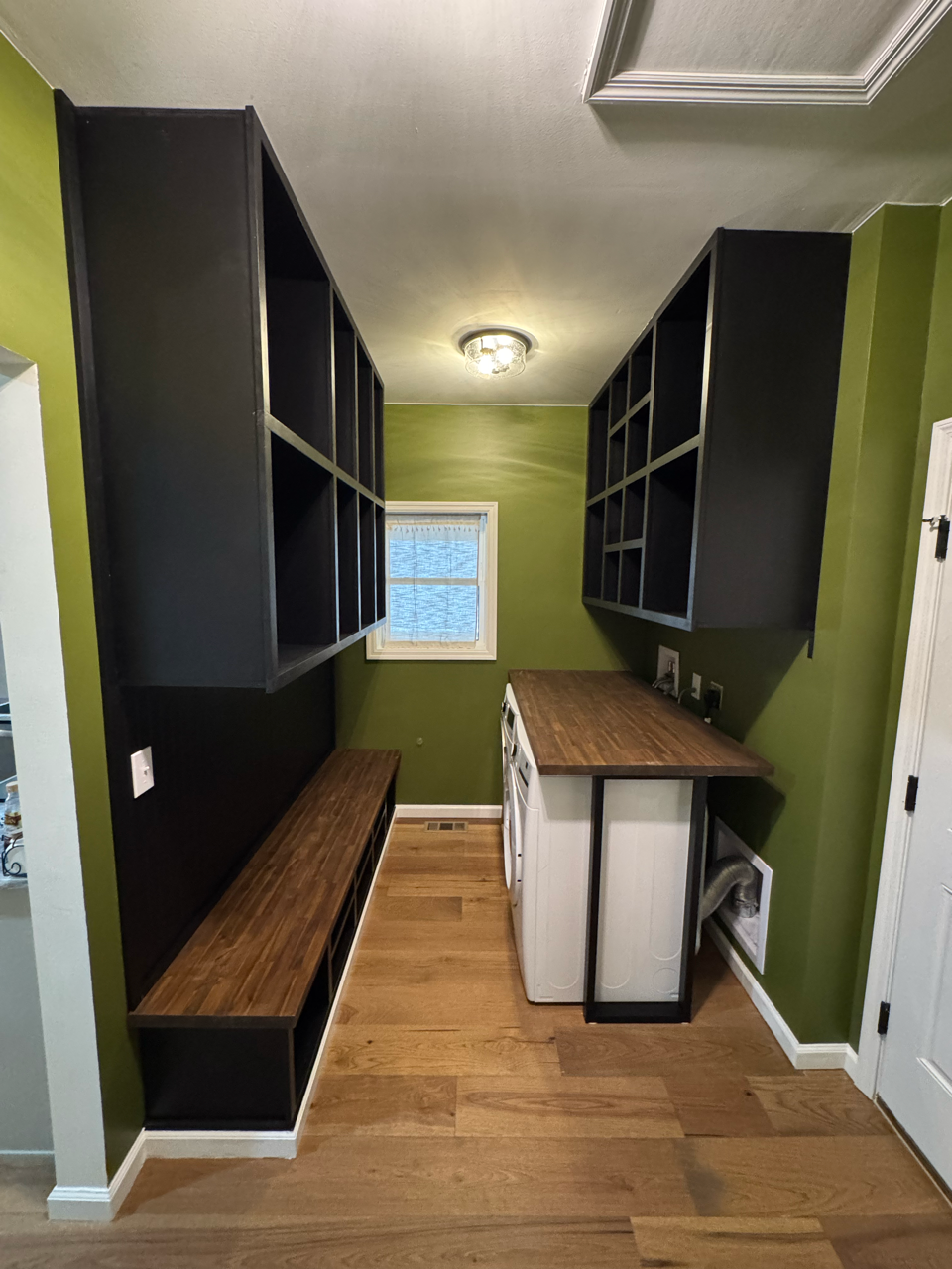 A laundry room with olive green walls, a wooden bench, wall-mounted storage units, and a wooden countertop over machines.