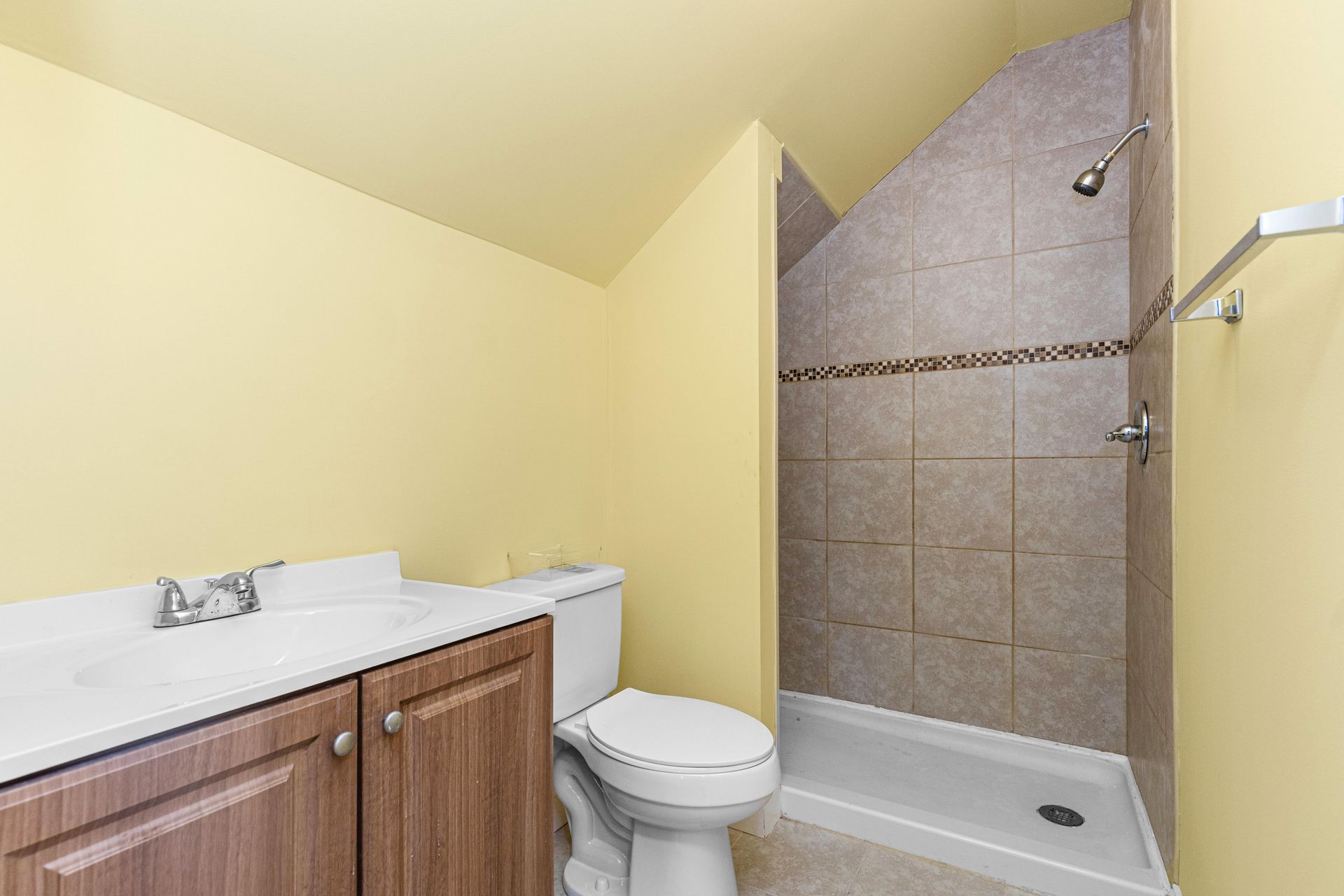 A bathroom with a wood-toned vanity, a white toilet, and a tiled shower stall against pale yellow walls.
