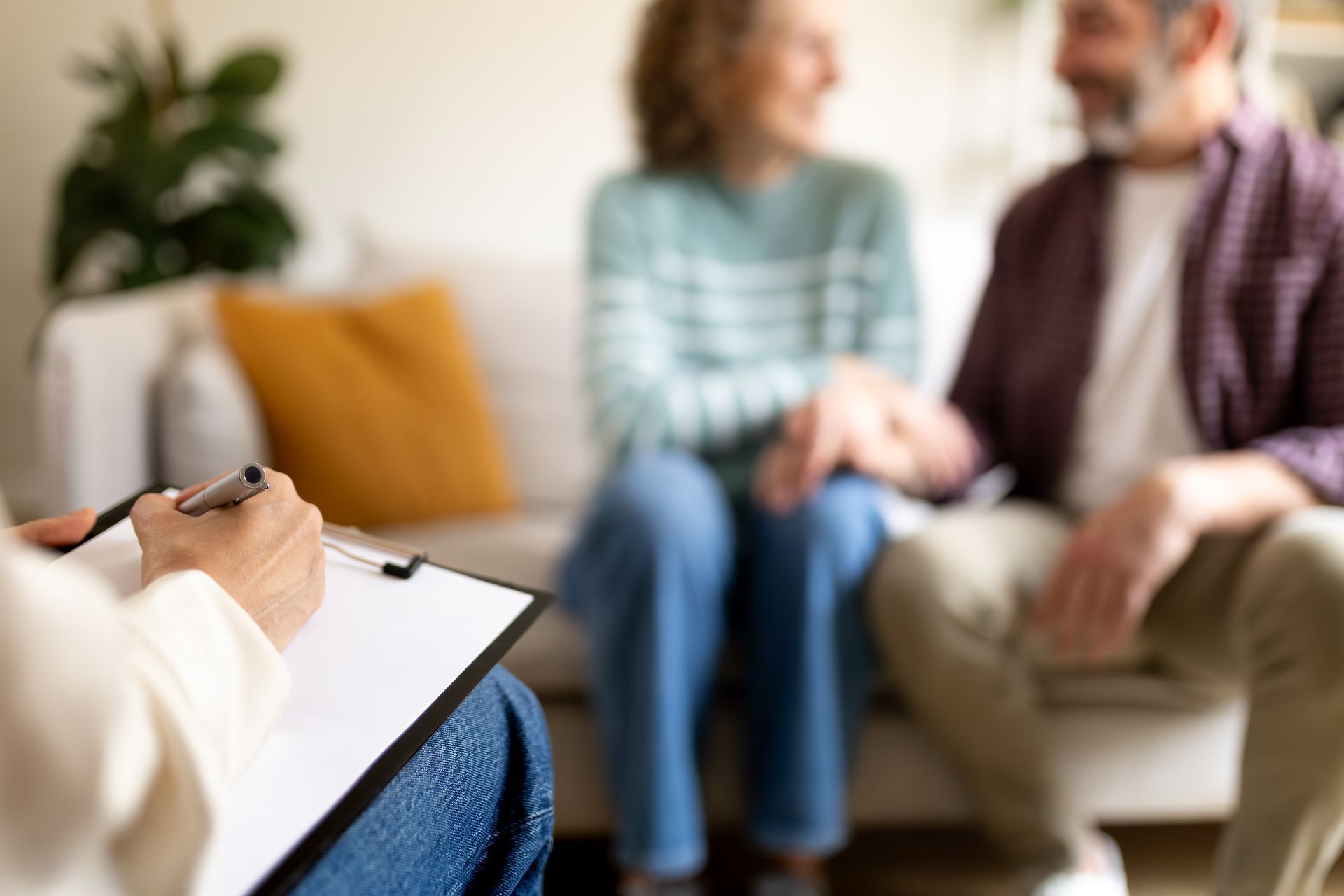 Therapist taking notes on a clipboard with a smiling couple blurred in the background.