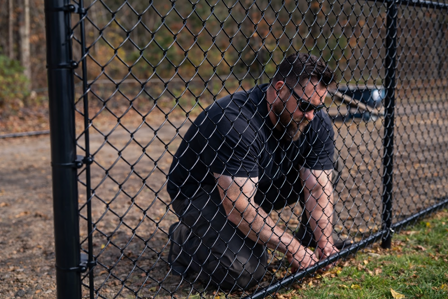 A person wearing sunglasses and a black shirt kneels by a black chain-link fence, working on the bottom edge.