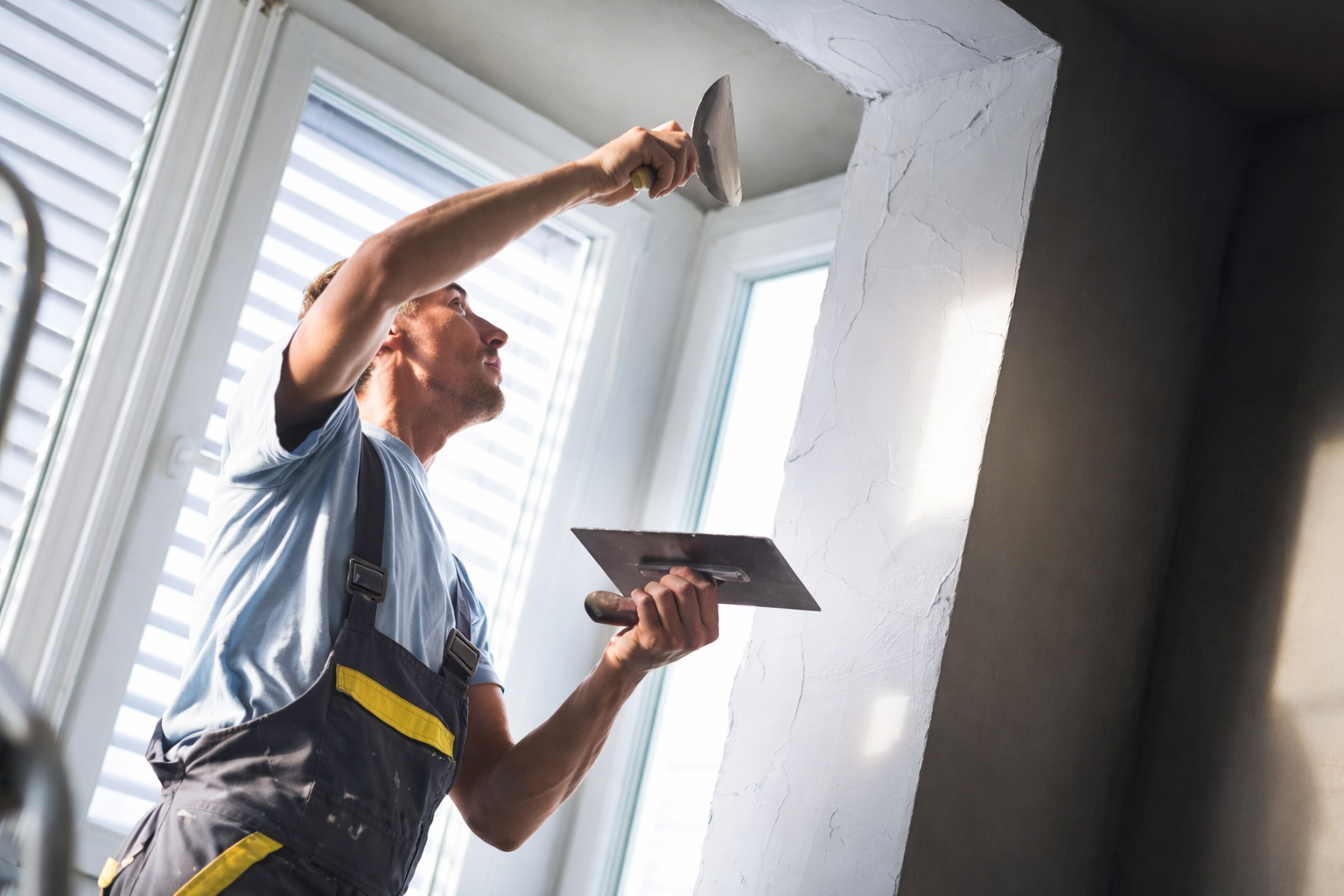 A construction worker wearing work overalls uses a trowel to apply plaster to a wall corner near a bright window.
