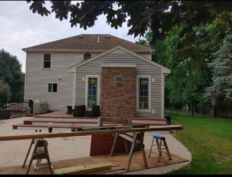 Back of a house with light siding and a stone accent, surrounded by greenery. Sawhorses and lumber in the foreground.