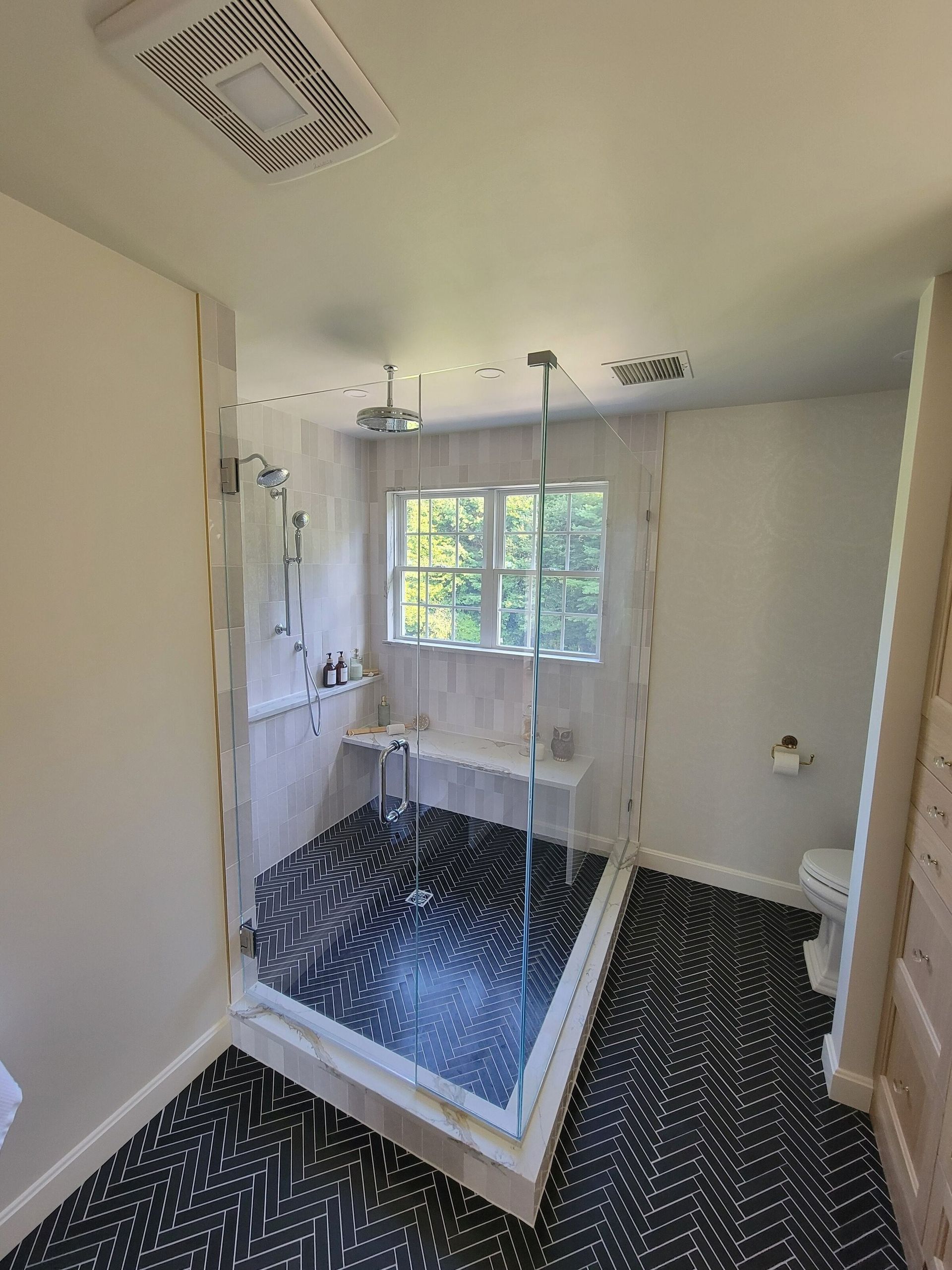 Modern bathroom with glass shower, black patterned floor, white walls, toilet, and window.