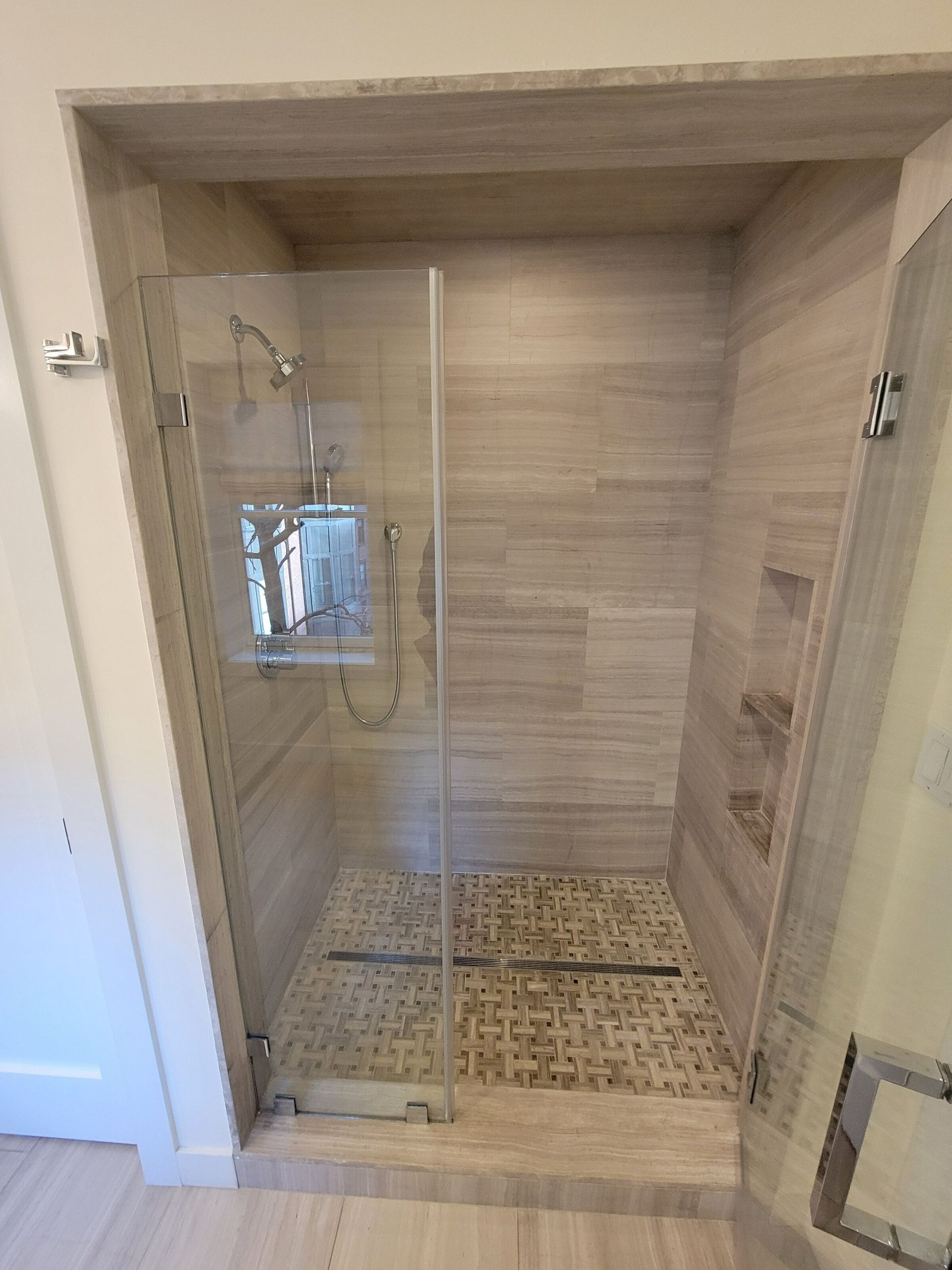 Shower stall with tan tiled walls and floor, glass door, and chrome fixtures.