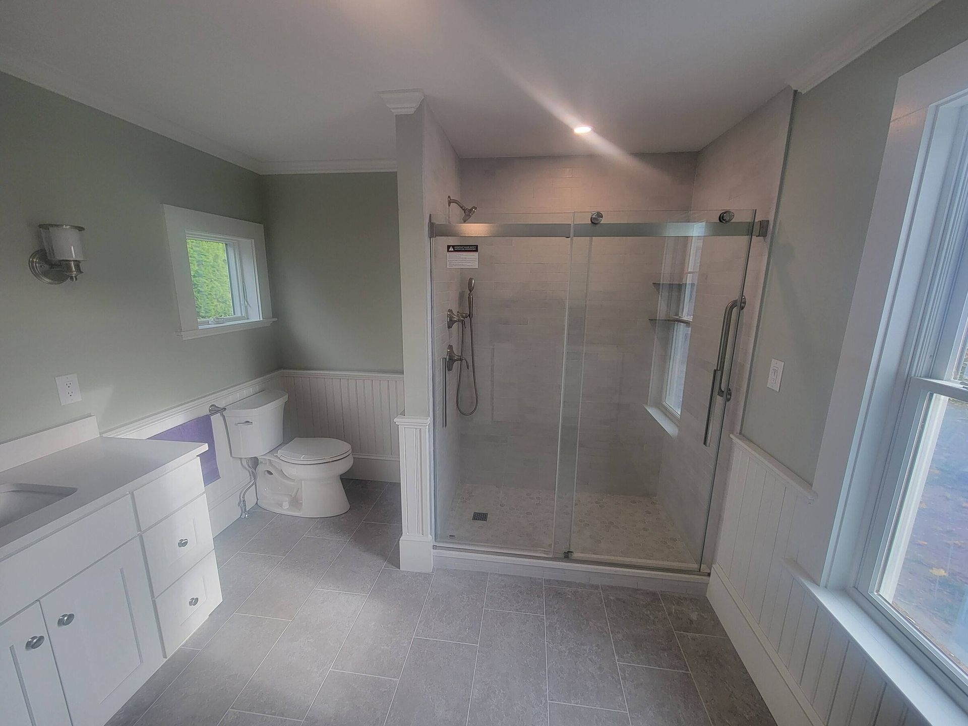 Bathroom with shower, toilet, and vanity. Gray tile floor, light green walls, white trim and cabinets, and glass shower doors.