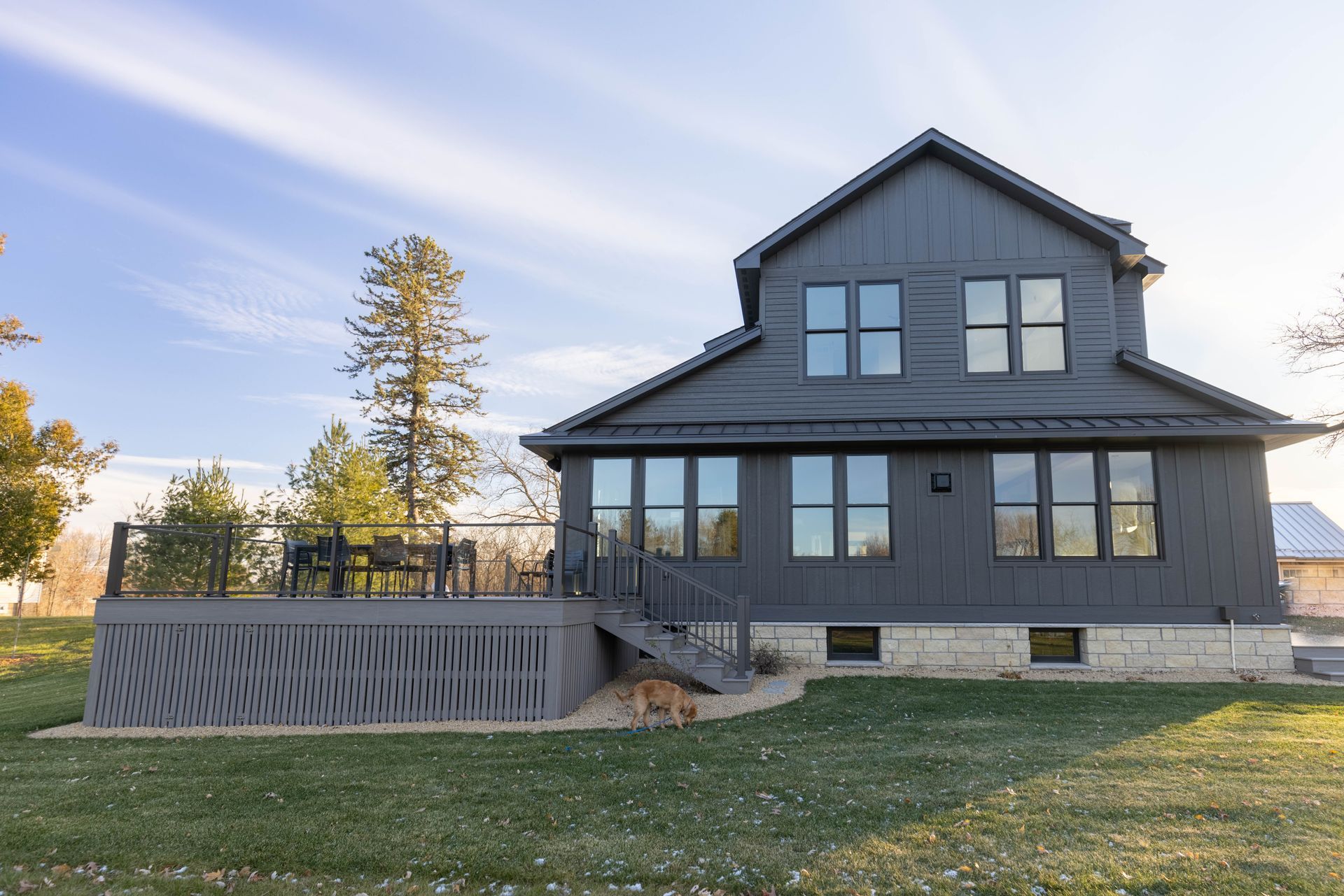 A large black house with a deck and stairs is sitting on top of a lush green field.