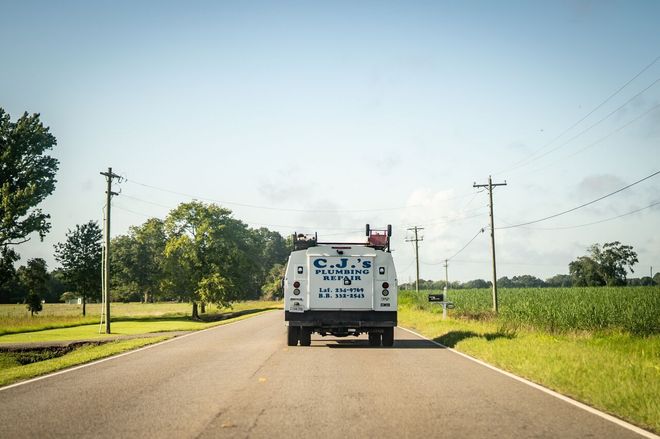 White utility truck driving on a rural road under a clear sky, with green fields on both sides.