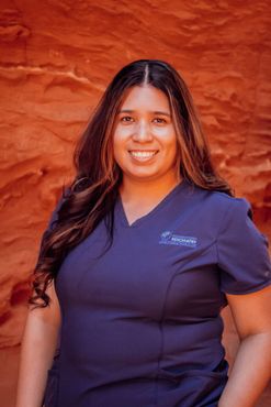 A woman in a blue scrub top is smiling in front of a red rock wall.