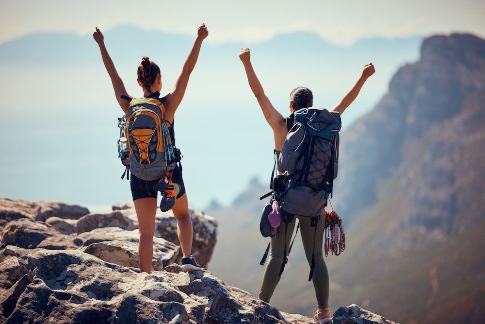 Two women with backpacks are standing on top of a mountain with their arms in the air.