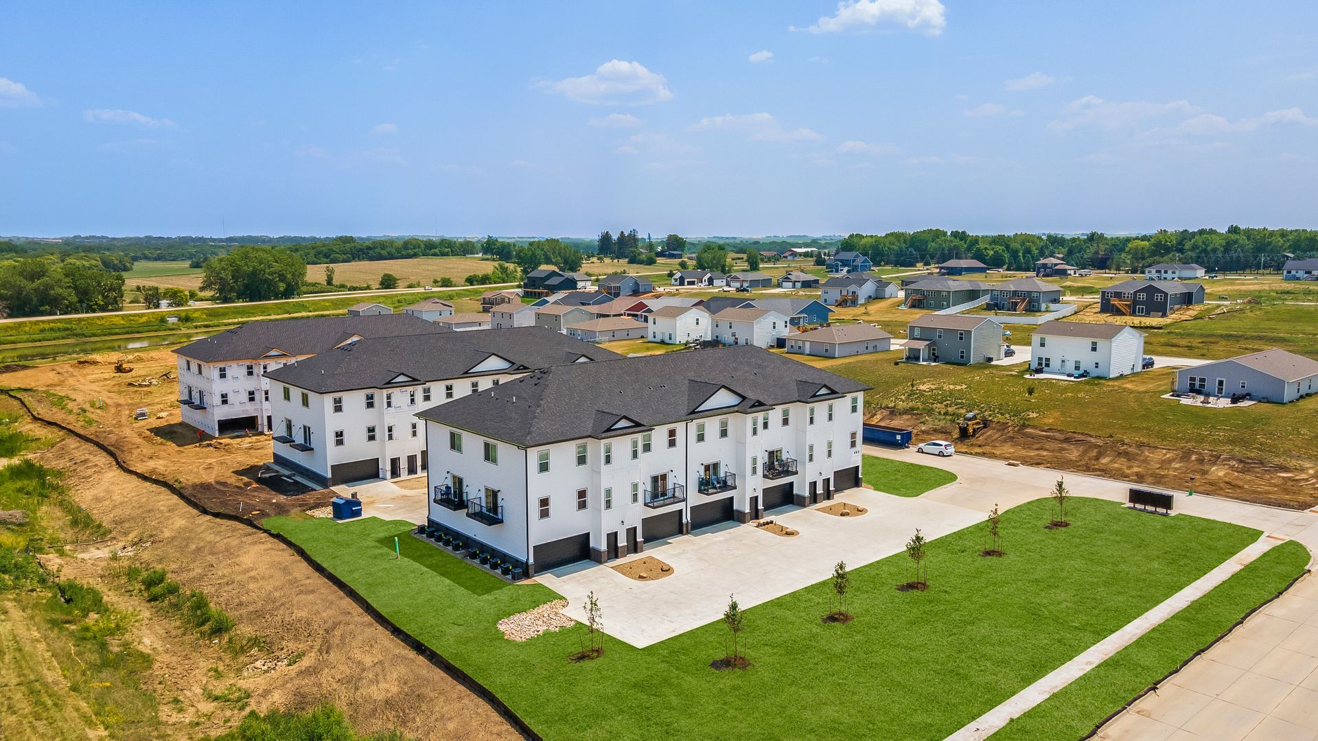 Aerial view of a new white townhouse complex with black roofs, surrounded by undeveloped land and a concrete driveway.