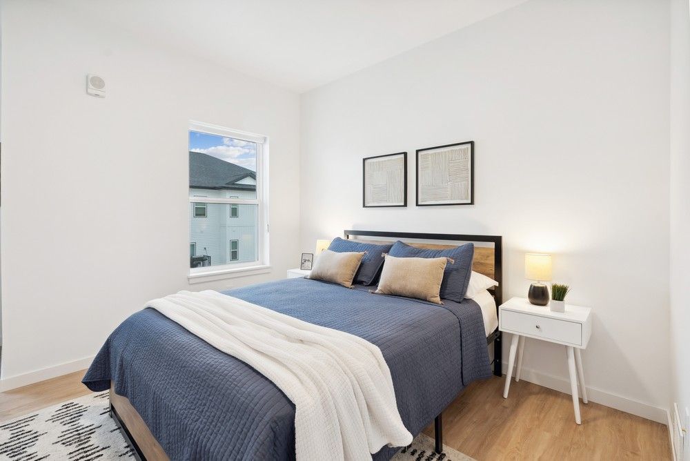 A bedroom with a blue quilt on the bed, two beige pillows, a white nightstand with a lamp, and wall art in a bright room.
