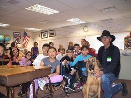 A man in a cowboy hat is kneeling next to a dog in front of a group of children in a classroom.