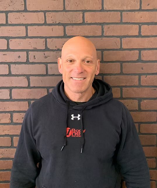 Bald man in a black hoodie with red logo smiles in front of a brick wall.