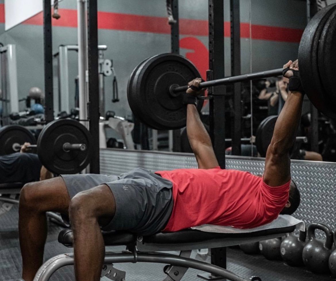 Woman in gym holding barbell, preparing to lift. Gray tank top, focused expression. Other people and equipment in background.