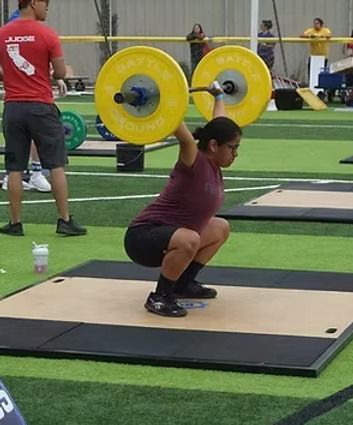 Weightlifter in deep squat, lifting barbell overhead. Yellow weights, green turf, judge in red shirt.