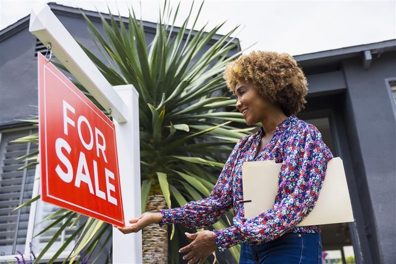 Person standing next to a 'For Sale' sign in front of a house.