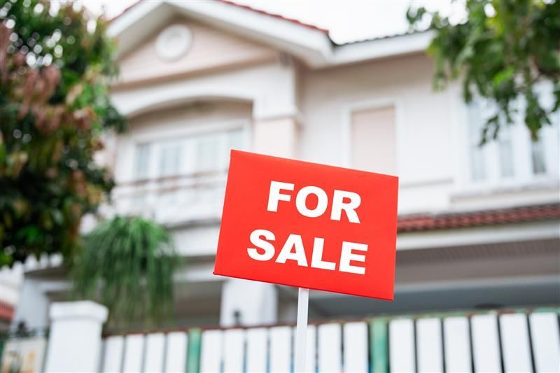 focused view of a red “for sale” sign outside a white house.