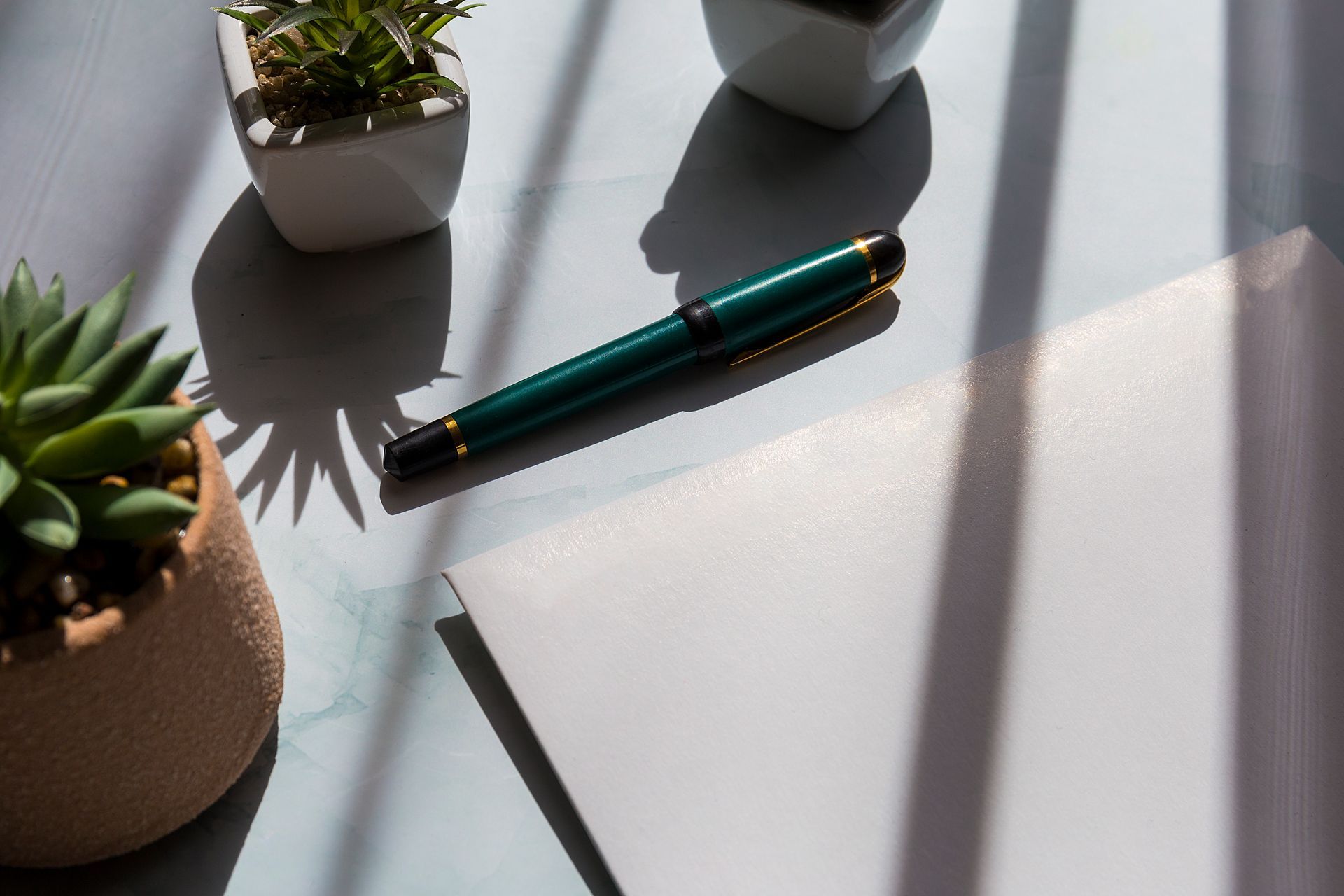 Green pen on white notebook, two small plants in pots. Sunlight and shadows.