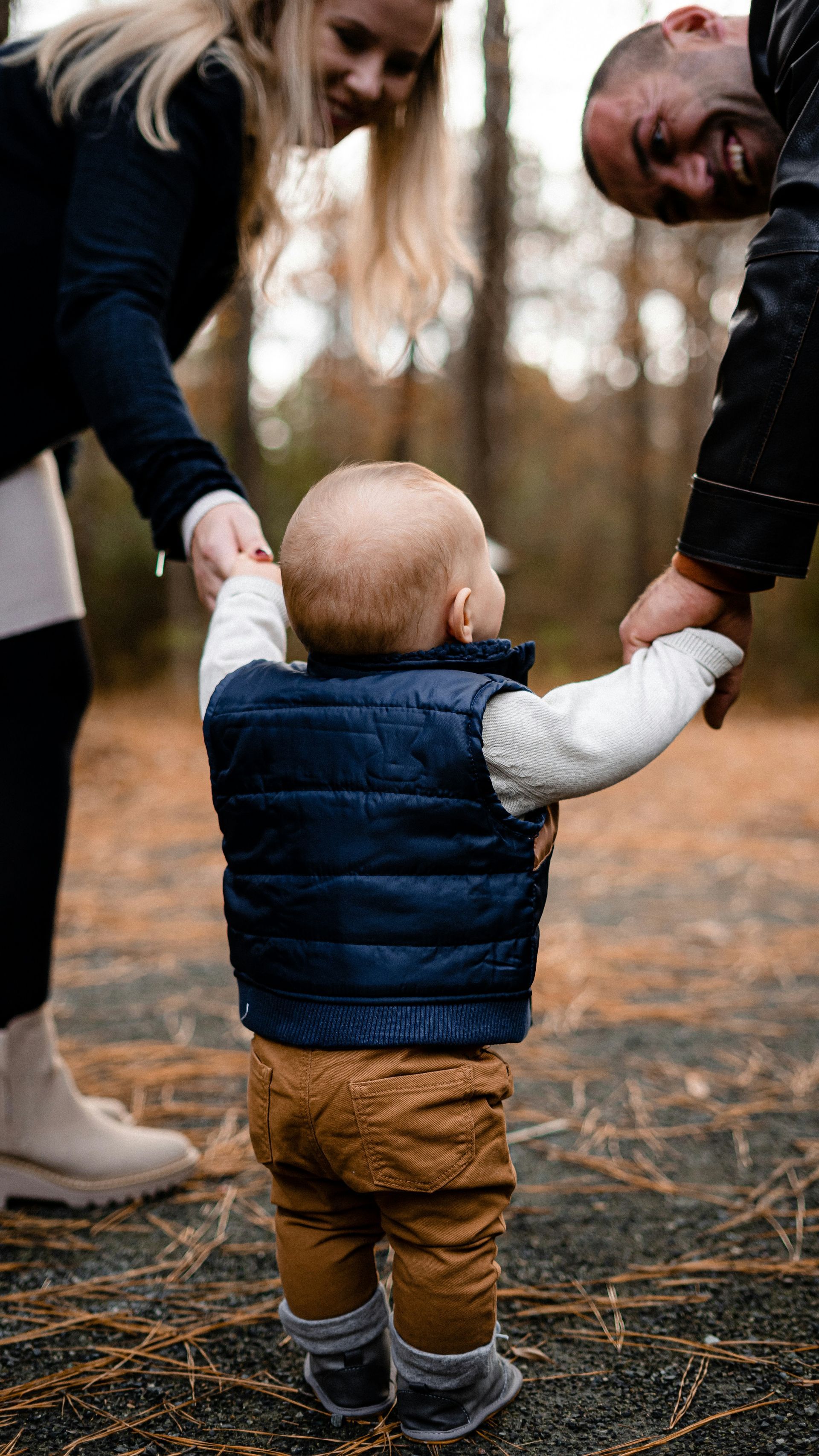 Parents helping a baby walk in a wooded area. Baby wearing a vest, holding hands.