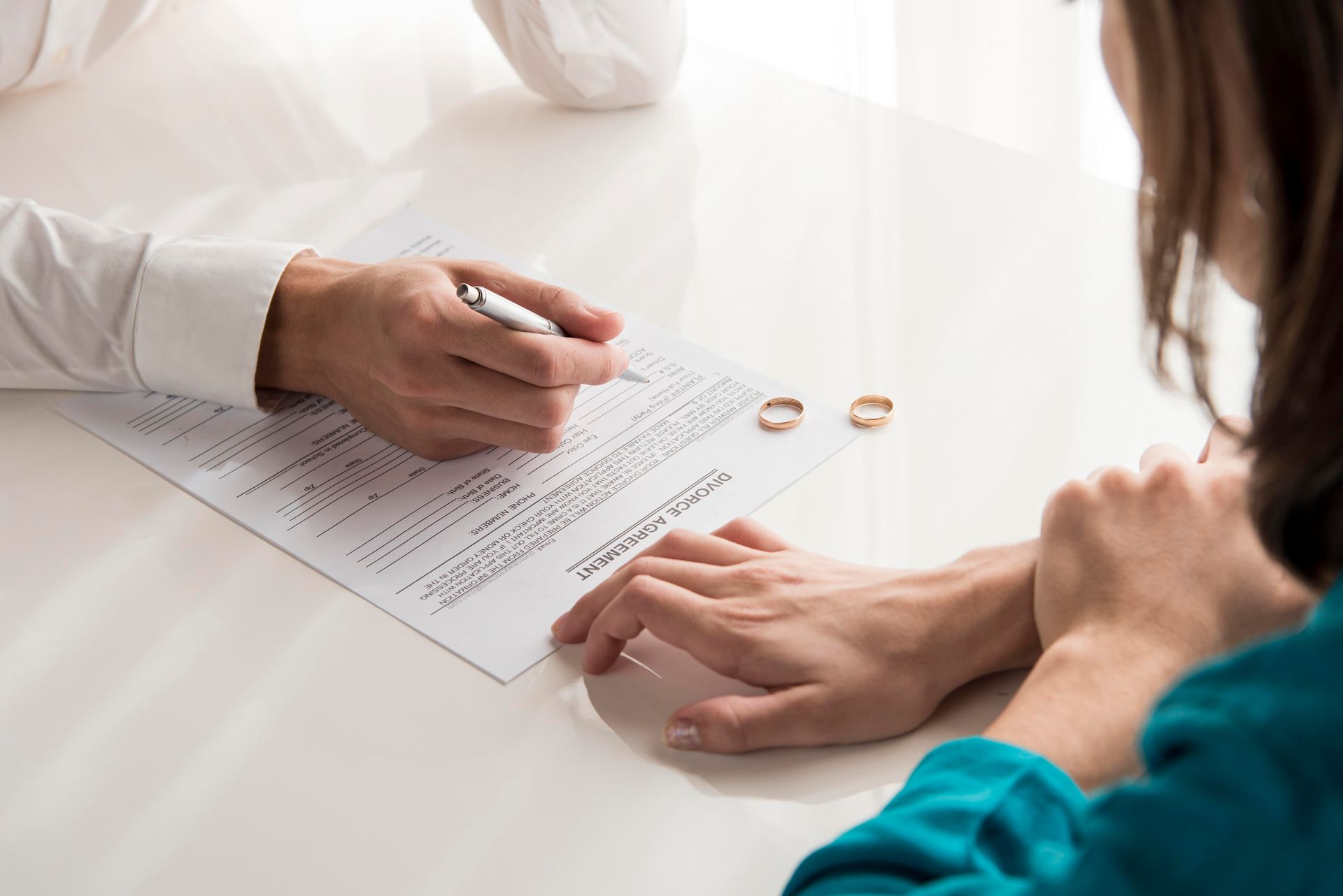 A person signing divorce papers at a table with rings; another person watches.