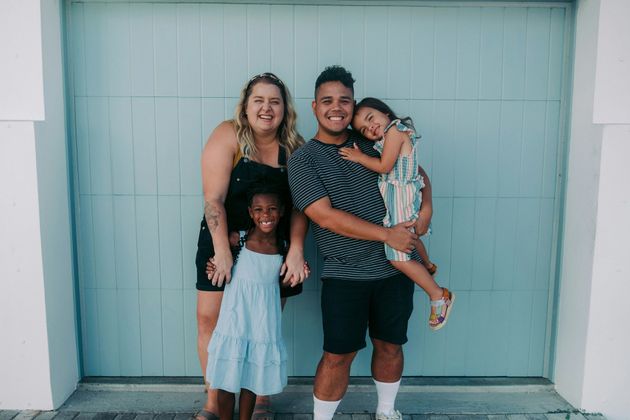 Family of four smiling in front of a light blue door: parents, a girl held, and a girl standing.