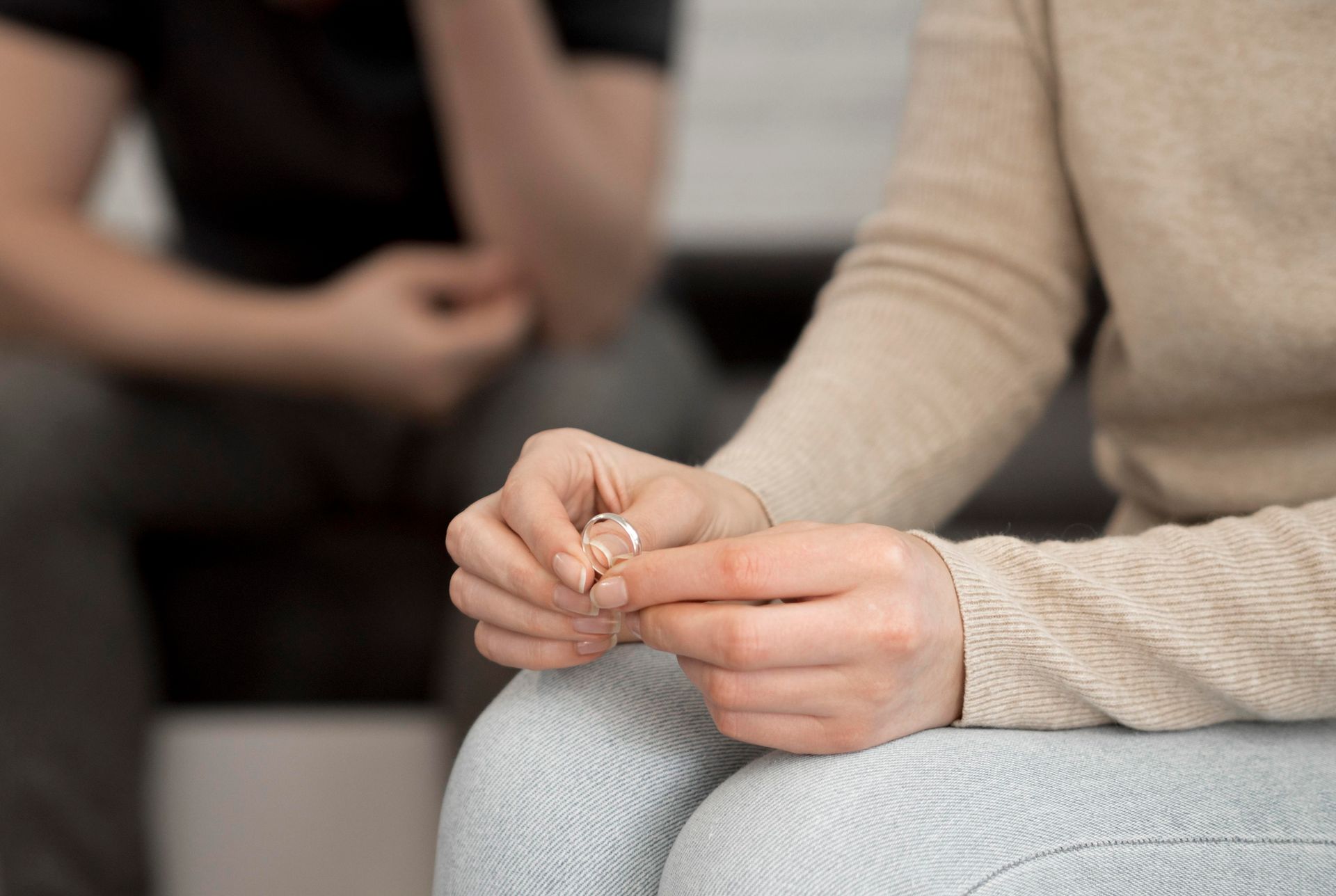 Woman holding wedding ring, man blurred in background, likely in a divorce setting.