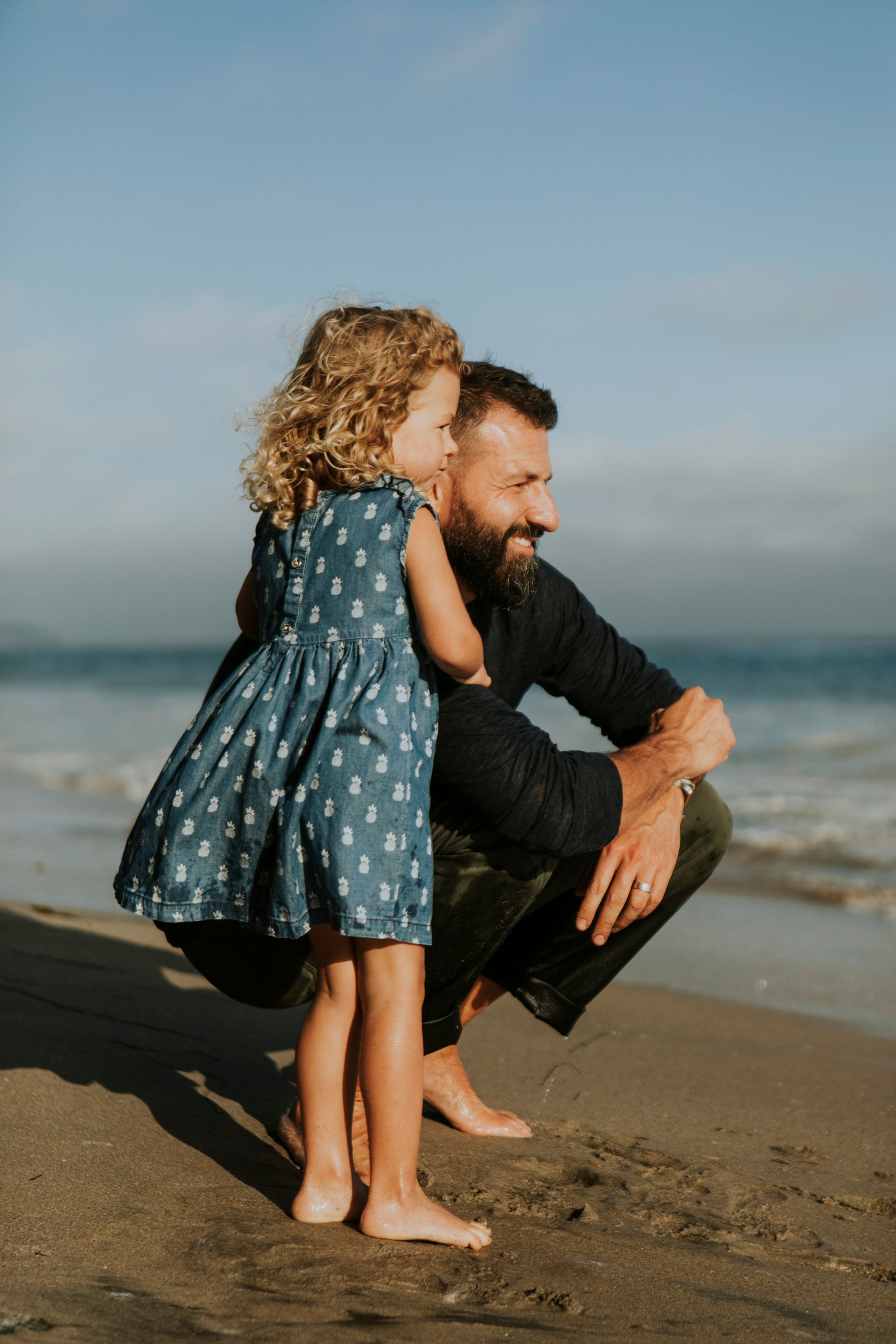 Father and daughter at beach, squatting, looking at ocean. Girl in blue dress, man with beard, blue sky.