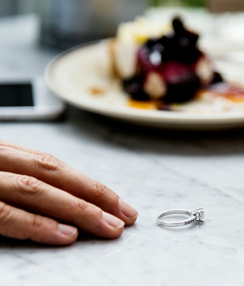 Hand reaching for a diamond ring on a marble table, with a blurred dessert plate in the background.