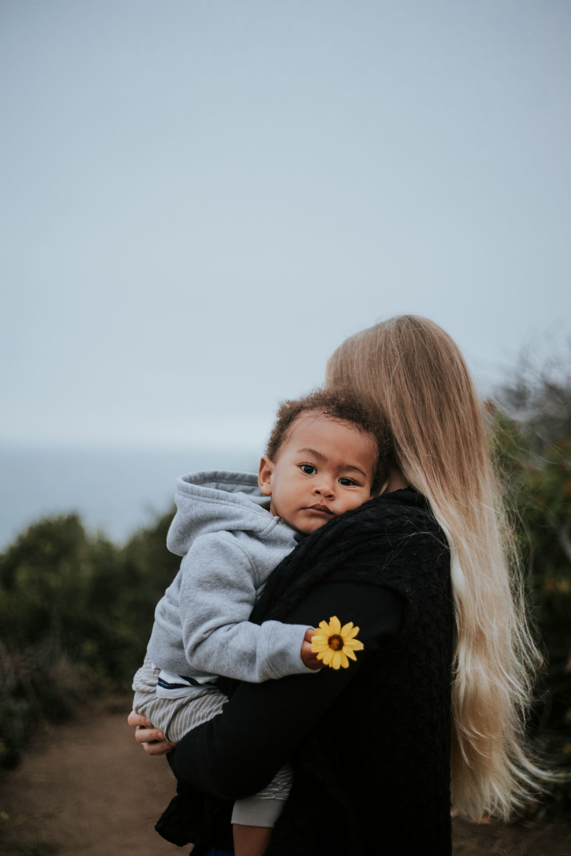 Woman holding a baby, both looking at the camera. Baby holds a flower, overlooking a cloudy ocean.