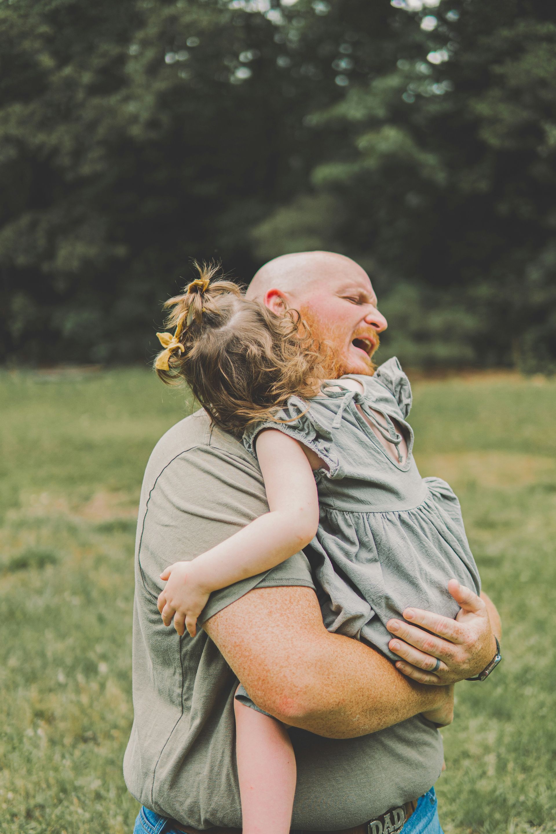 A man with a shaved head laughing, hugging a young girl in a field. They're both smiling, enjoying a moment together.