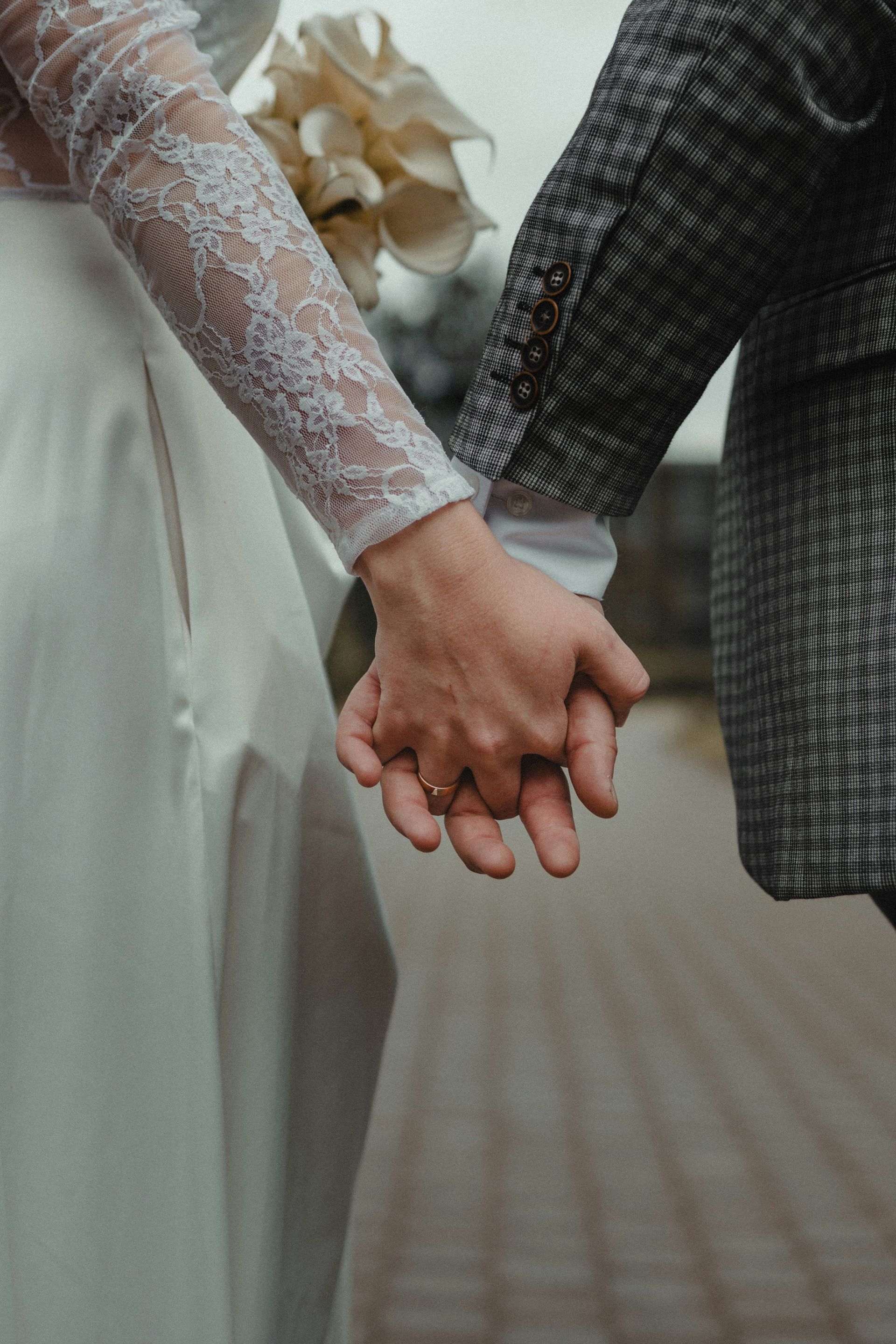 Bride in lace sleeve holds groom's hand, walking outside.