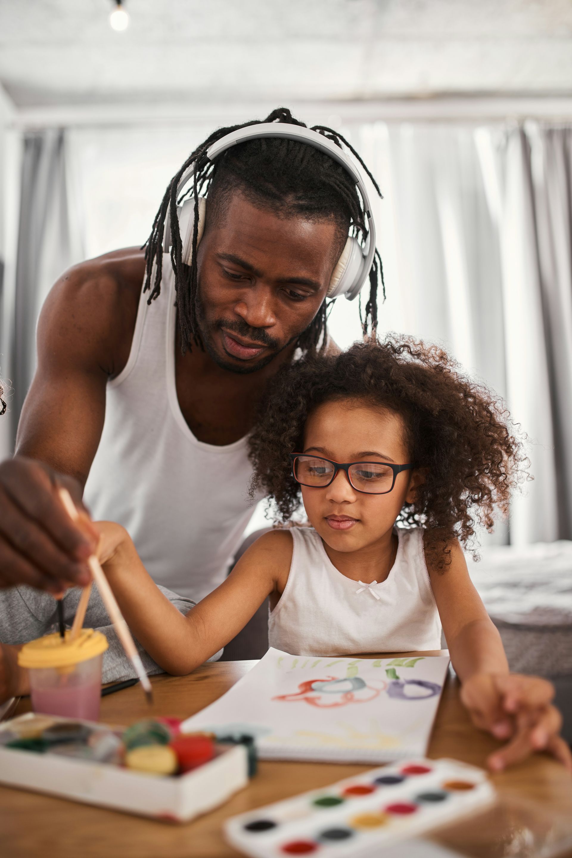 Father helps daughter paint with watercolors at a table; both wear white shirts, she wears glasses.