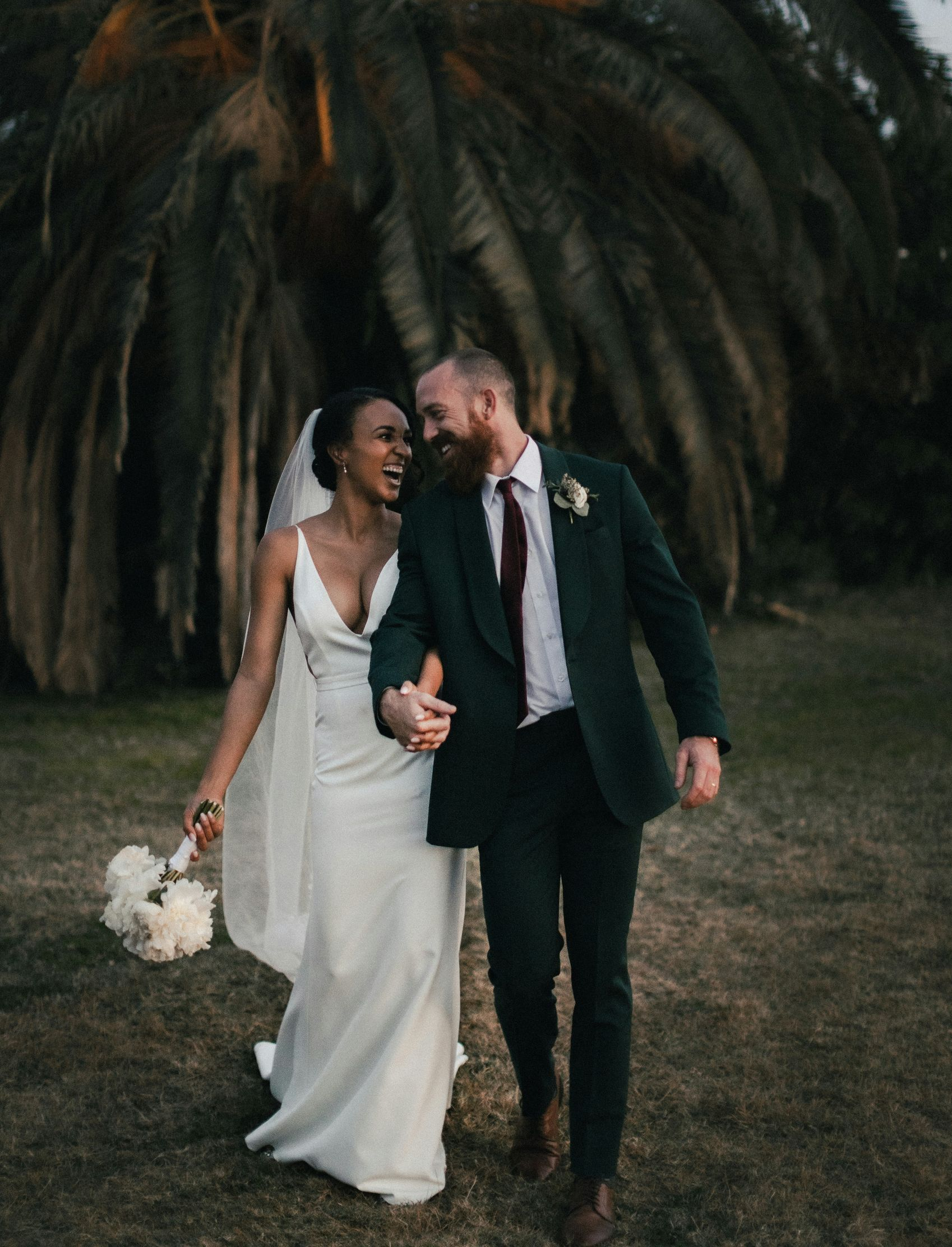 Newlyweds walking arm in arm, smiling; bride in white dress, holding flowers; groom in green suit, outdoor setting.