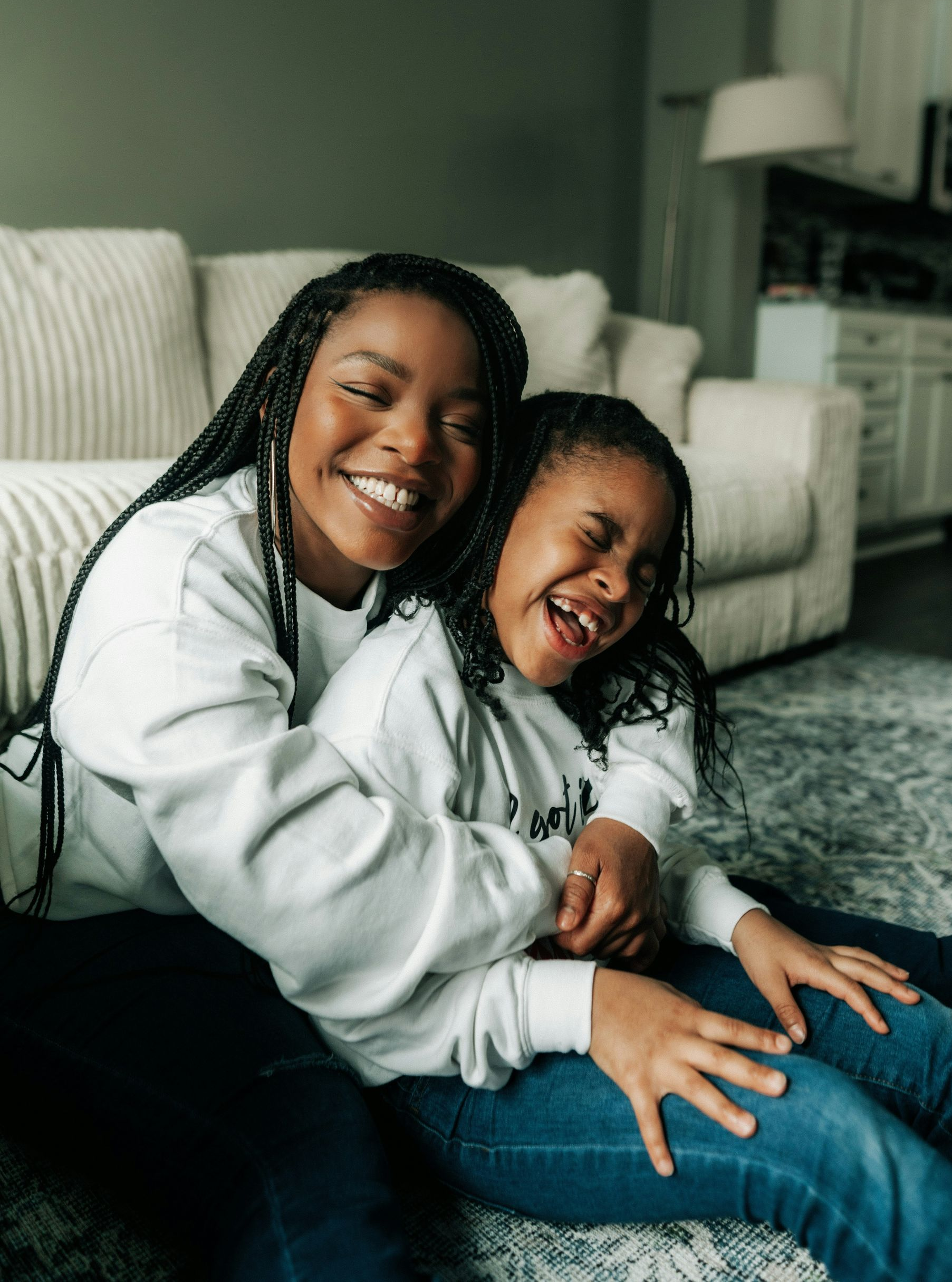 Two smiling Black women hugging on a floor. One is older with braids; they wear white sweatshirts and jeans.