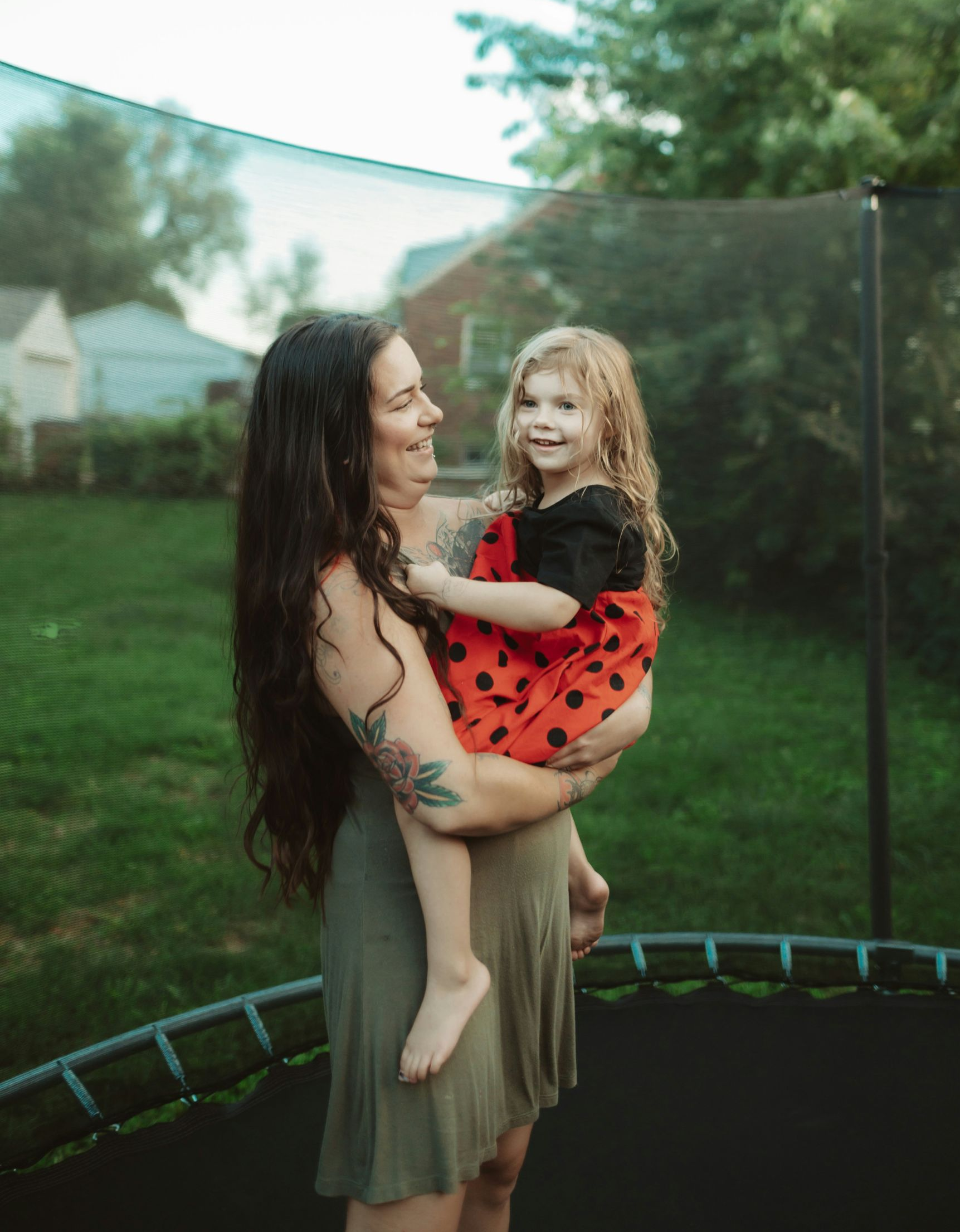 Woman holding a smiling child on a trampoline in a backyard. Green dress, red dress.